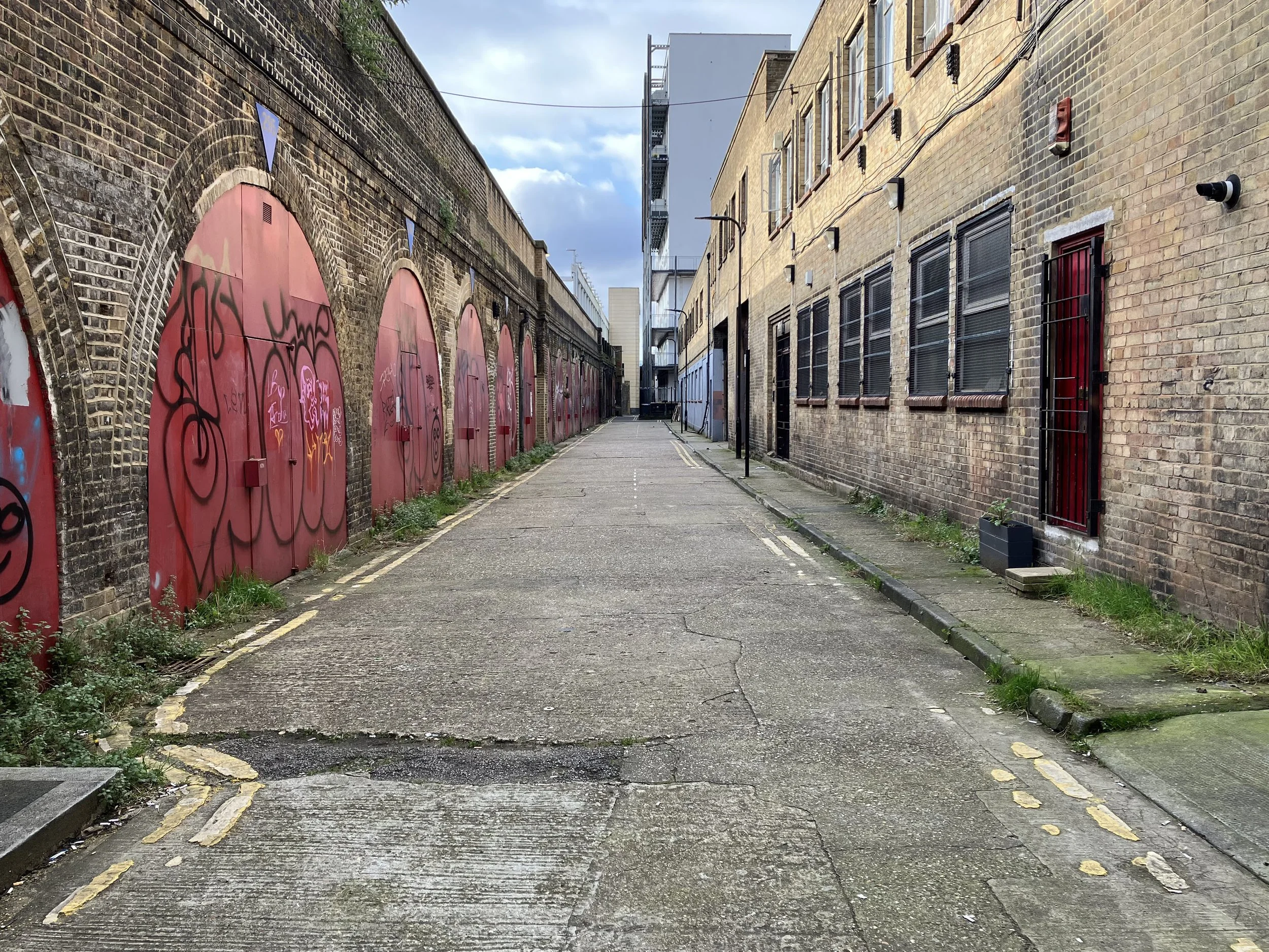 An empty graffiti-covered alleyway between brick buildings with a cloudy sky overhead.