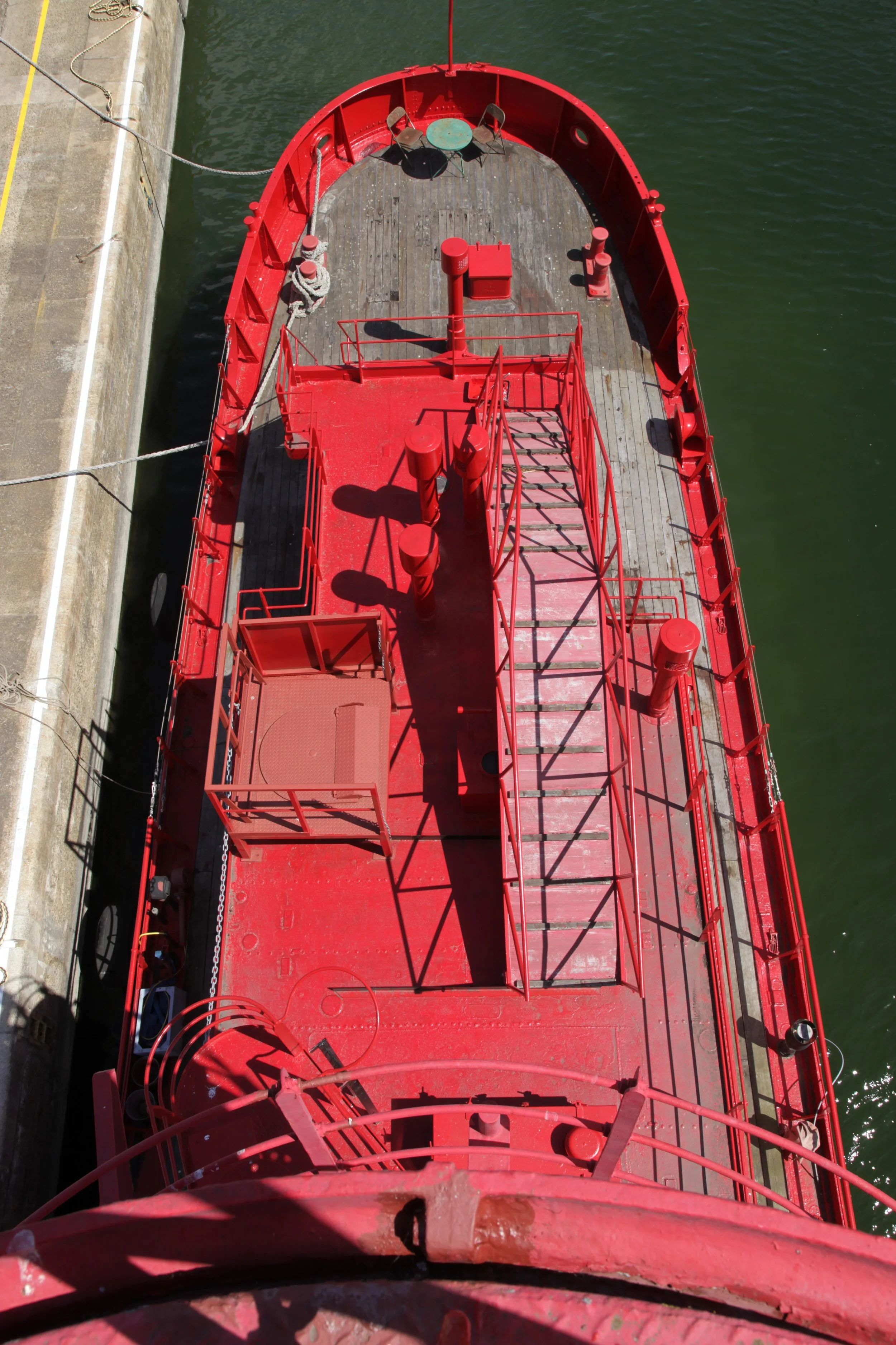 Top view of a red ship docked at a pier, with a wooden deck, railings, and equipment on the deck.