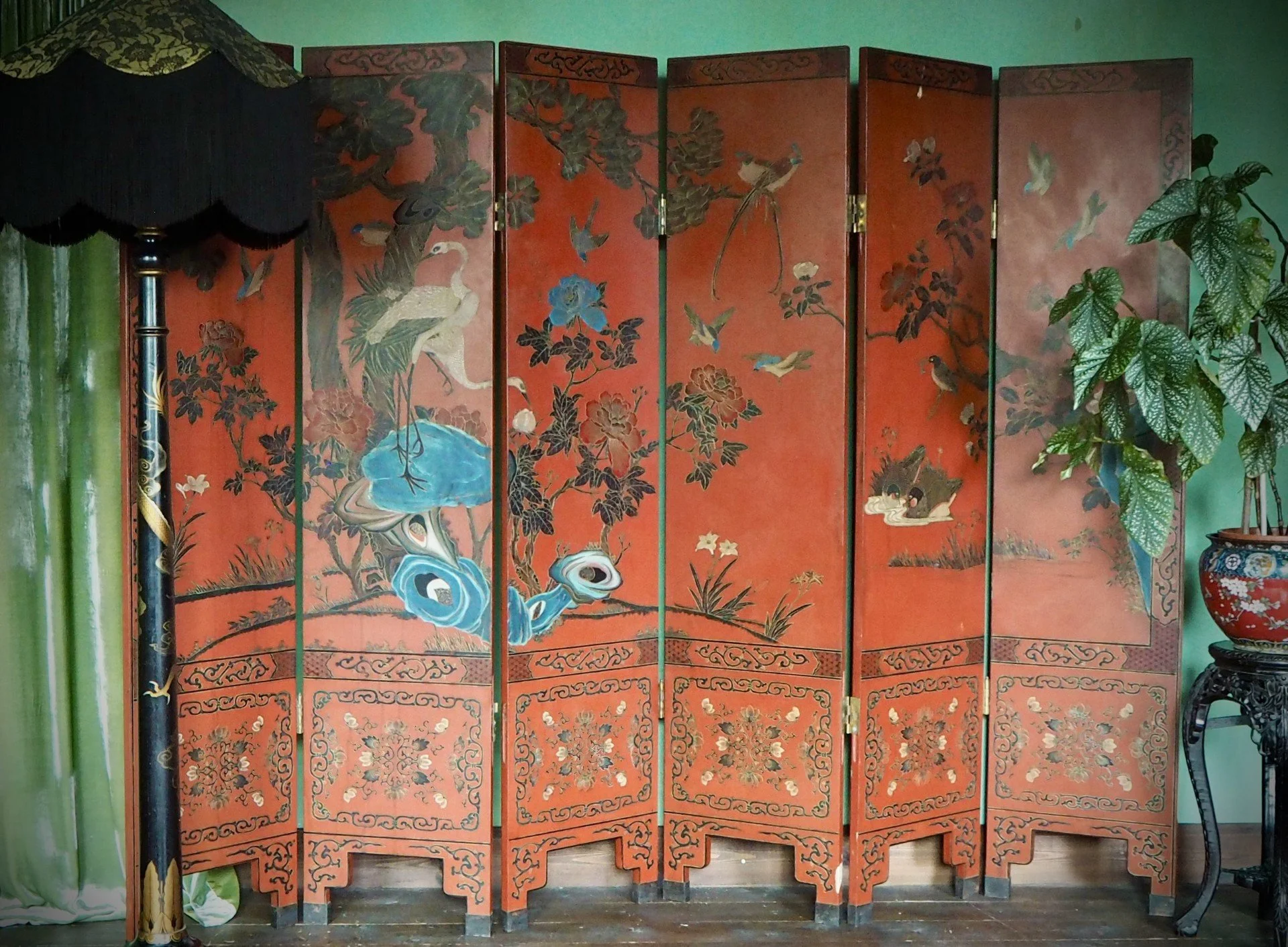A decorative folding screen with Asian-style painting of a crane, flowers, and birds on an orange background, placed indoors next to green plants and a black side table.
