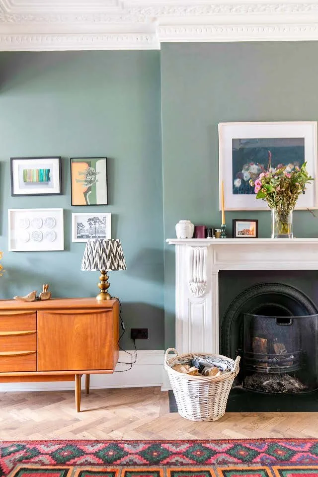 Living room corner featuring a green wall with framed artwork, a mid-century wooden sideboard, a patterned table lamp, a fireplace with a white mantel, a vase of flowers, and a basket with firewood, a red patterned rug on a wooden floor.