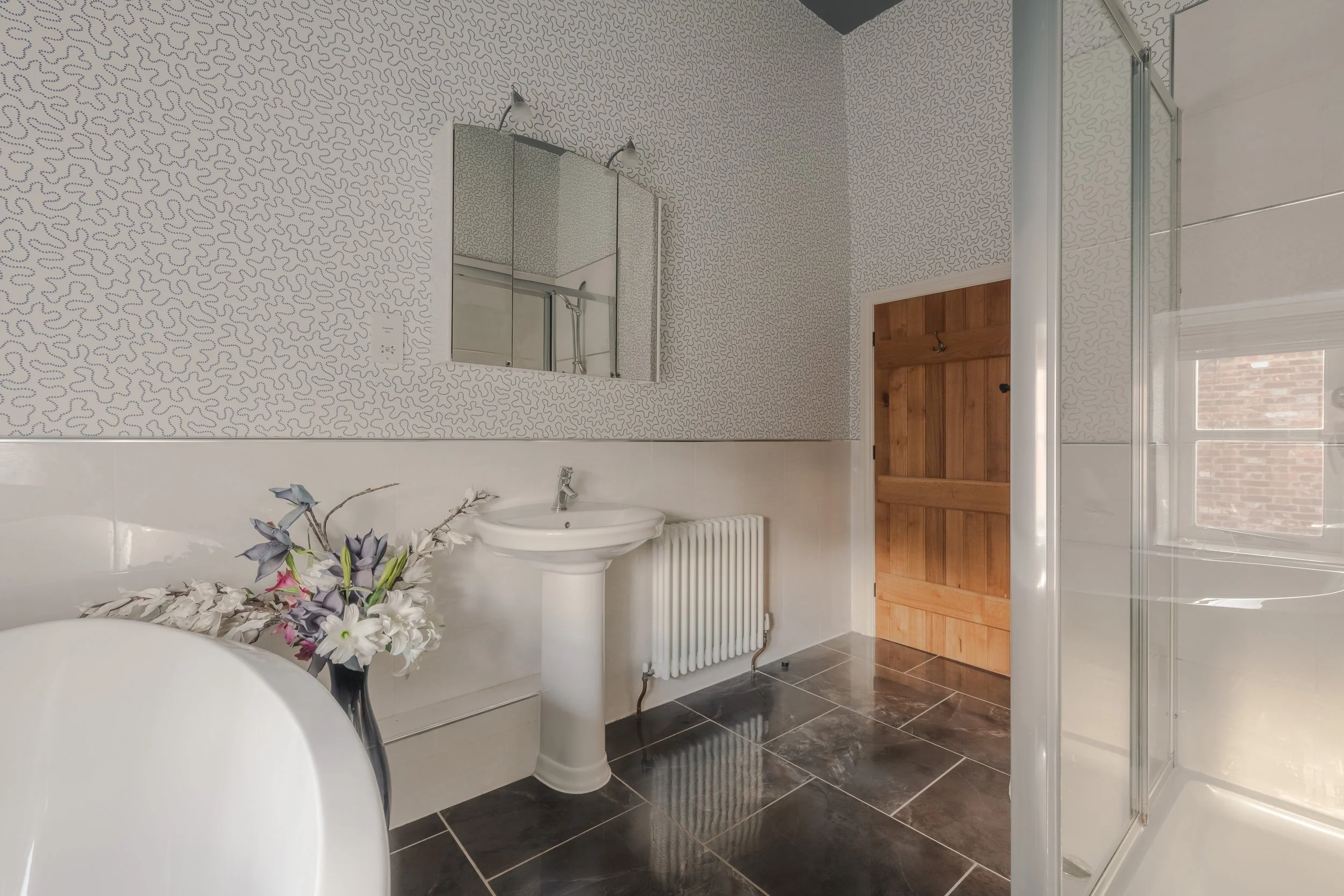 Bathroom with patterned wallpaper, white sink, mirror, radiator, wooden door, shower stall, and window with brick wall outside.