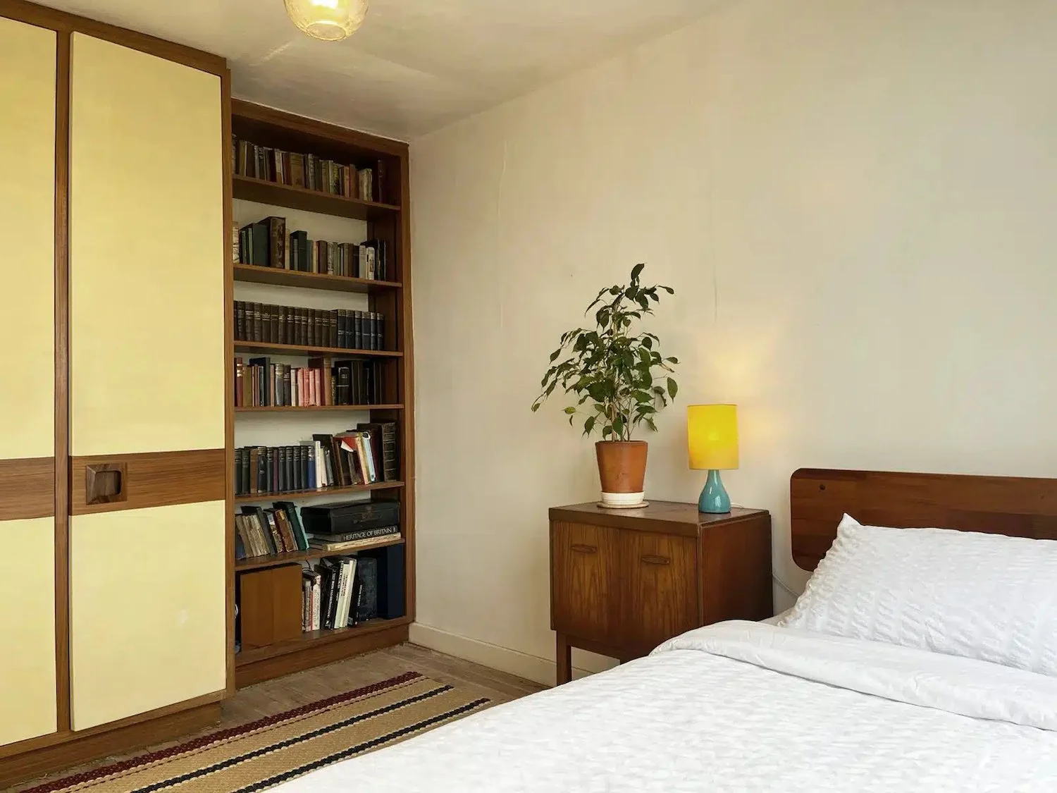 A bedroom with a wooden bed frame and white bedding, a wooden nightstand with a yellow and blue lamp, a potted plant, and a built-in bookshelf filled with books, a folding door with yellow panels, and a small rug on the floor.