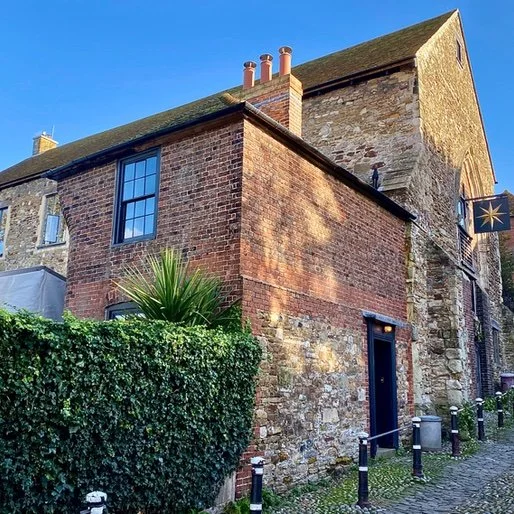 A historic brick and stone building with a sloped roof, a window, and a narrow black door, surrounded by a hedge and black posts with chains, with a bright blue sky in the background.