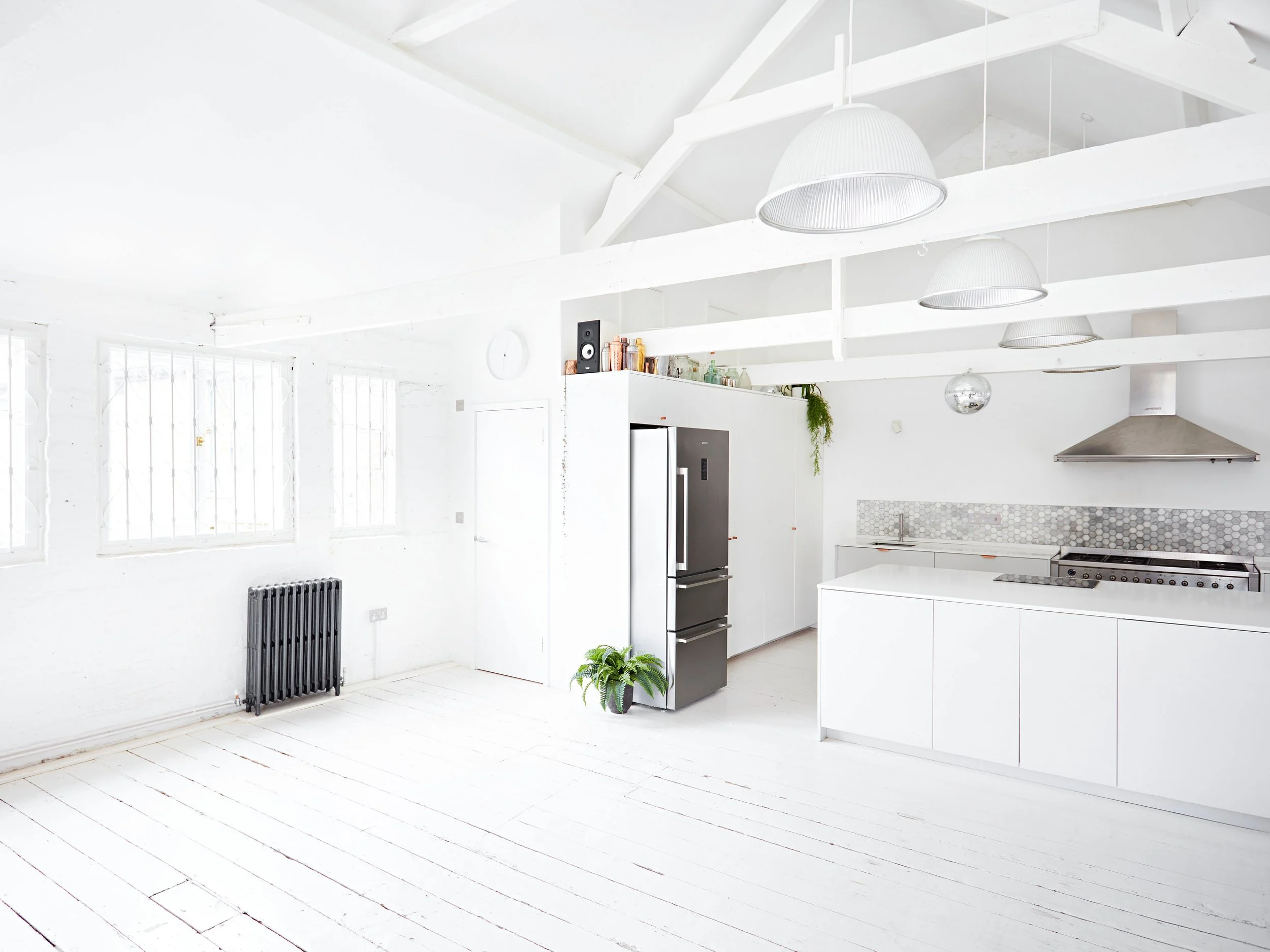 Bright, modern kitchen with white walls, ceiling, and flooring, featuring a stainless steel refrigerator, black plants, and minimal decor.
