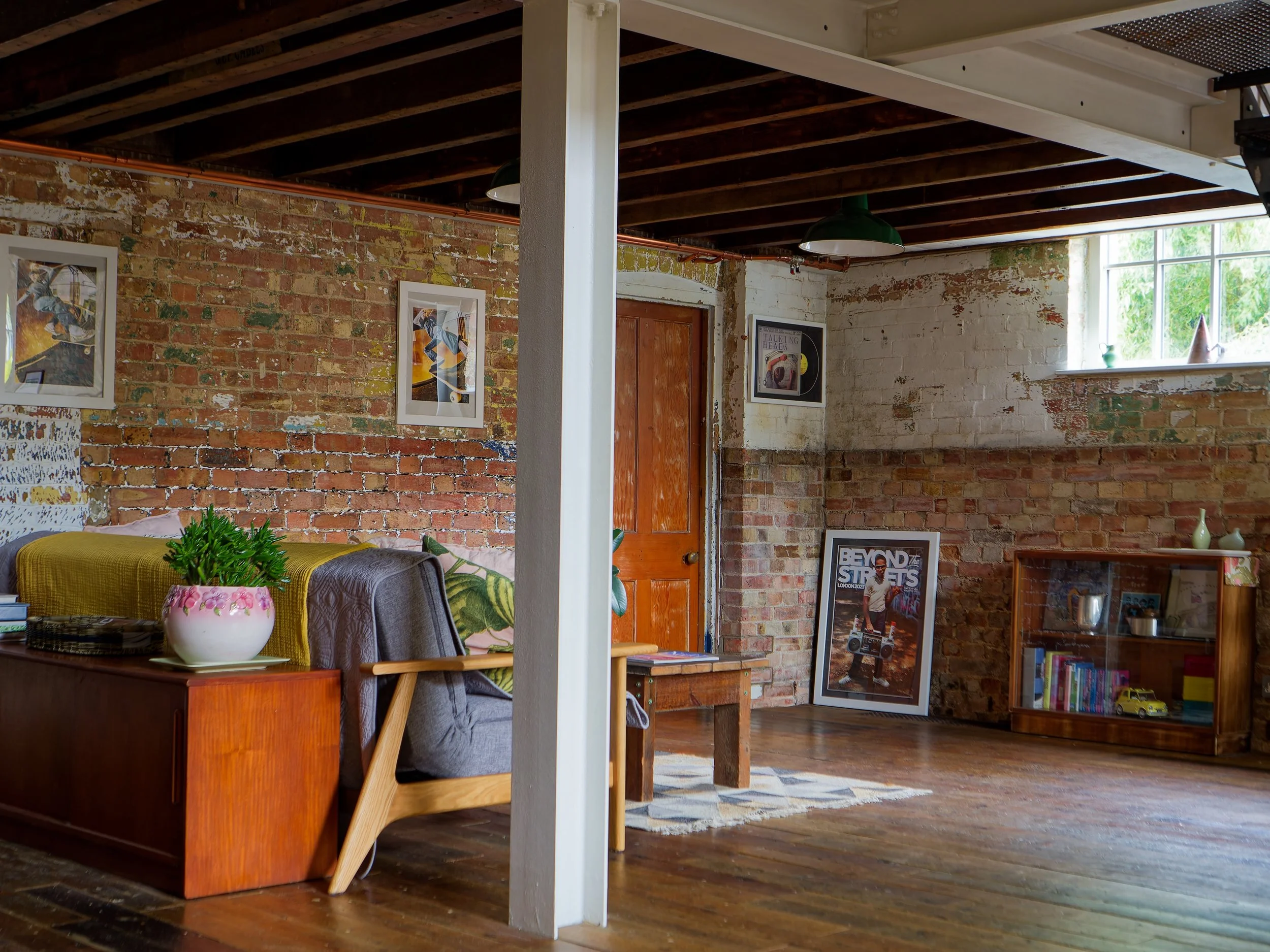 Interior of a loft-style living room with exposed brick walls, wooden ceiling beams, a window with sunlight, framed artwork, a wooden cabinet with books and decorative items, a framed magazine cover on the floor, and a couch with cushions.