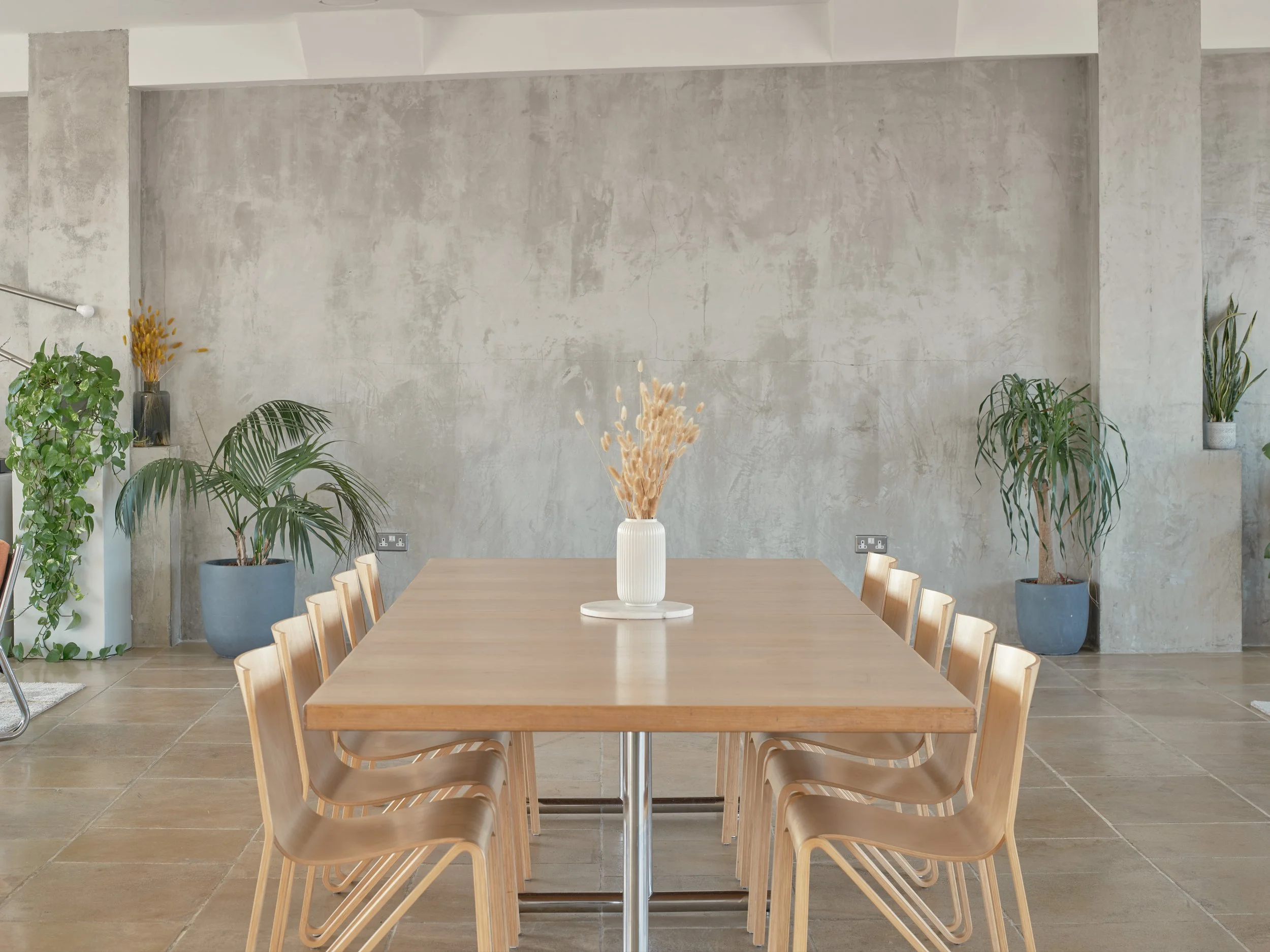 A minimalist dining room with a wooden table, surrounded by nine matching chairs. A white vase with dried flowers is centered on the table. The room has a concrete wall with smallsof potted plants on a wall shelf and the floor is tiled in large beige