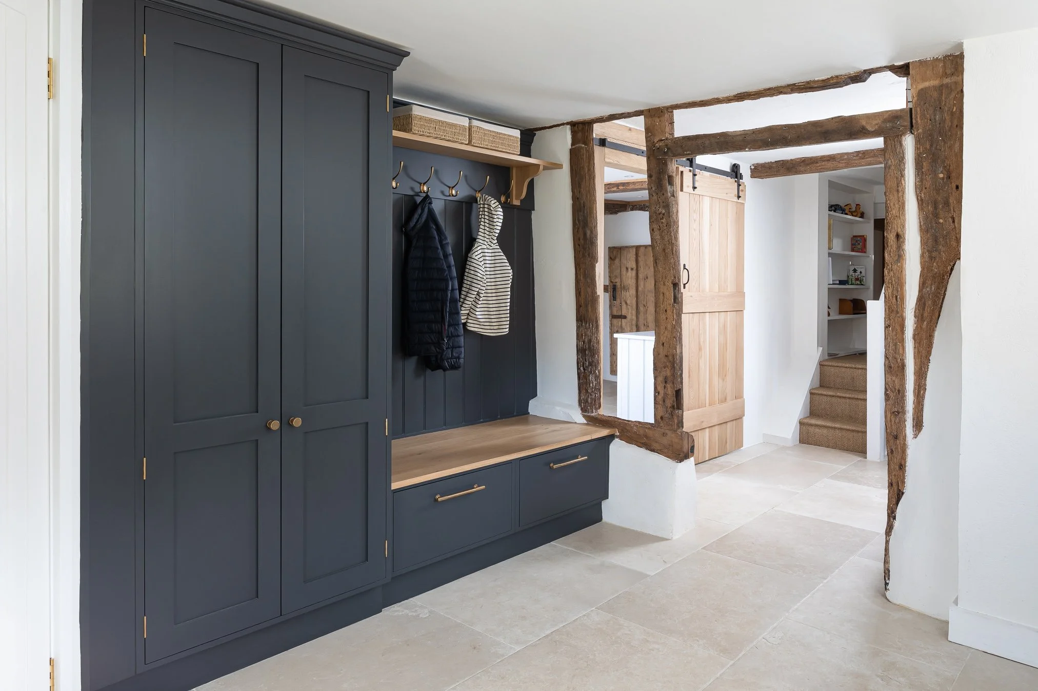Interior entryway with a dark blue built-in cabinet, hooks and a bench, exposed rustic wooden beams, a sliding barn door, stairs with carpet, and a white bookshelf in the background.