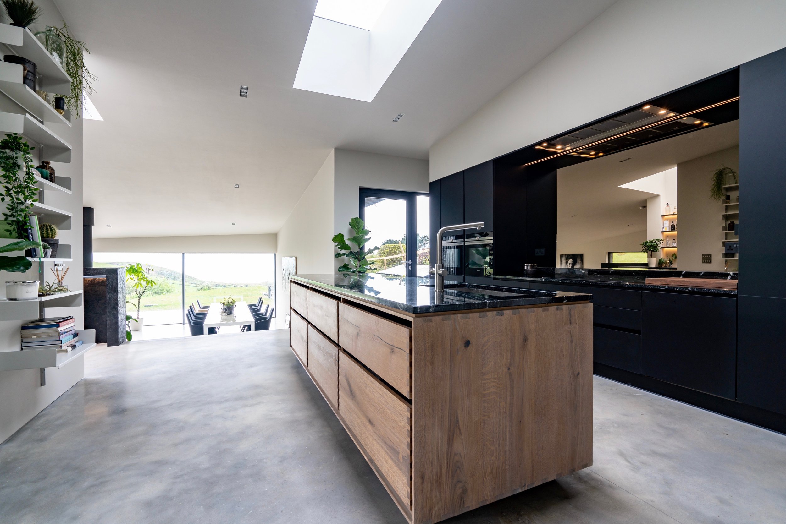 Modern open-concept kitchen and dining area with large windows and skylights, featuring black cabinetry, a wooden island with black countertop, and a view of green landscape outside.