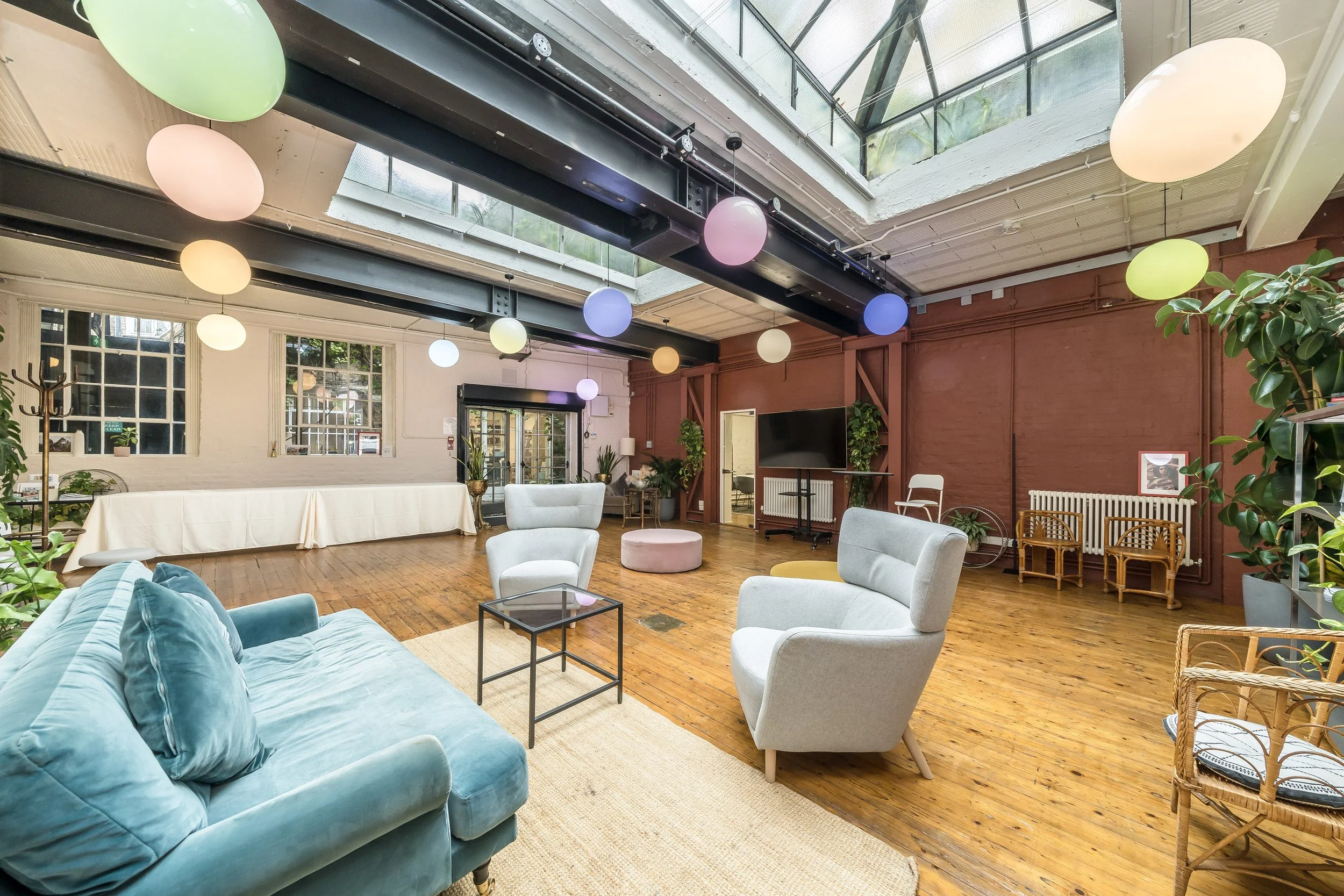 A spacious indoor lounge area decorated with multicolored hanging paper lanterns, featuring a teal velvet sofa, white armchairs, a flat-screen TV, potted plants, and large windows with a glass ceiling allowing natural light.