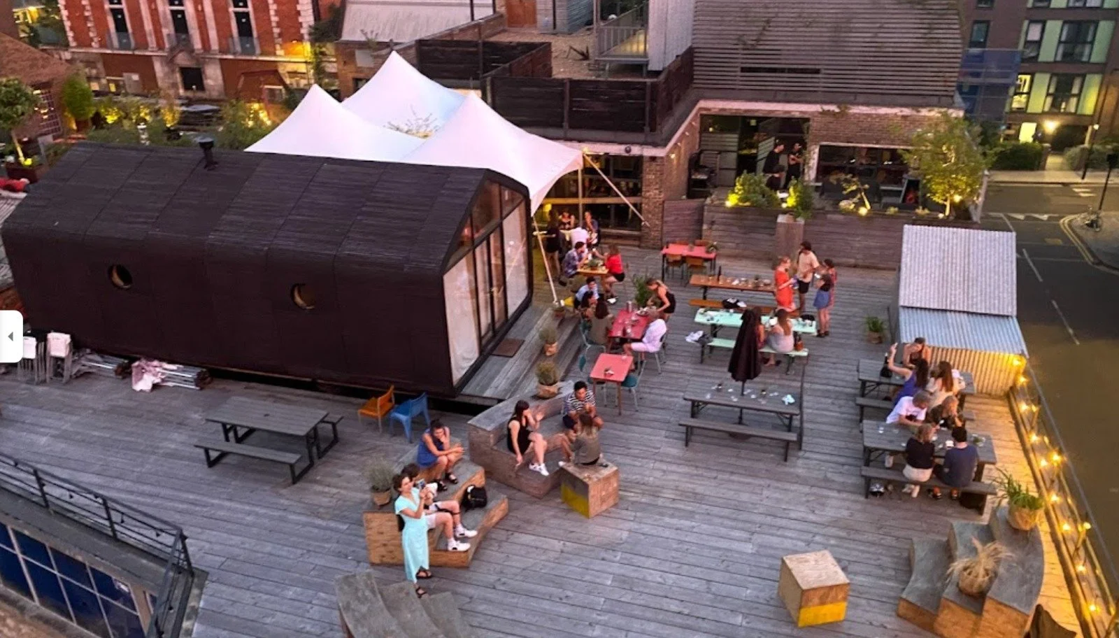 People gathered on a rooftop patio at dusk, seated at tables and benches, socializing and enjoying the evening. The space features wooden flooring, string lights along the railing, a small black building, and potted plants, with surrounding buildings