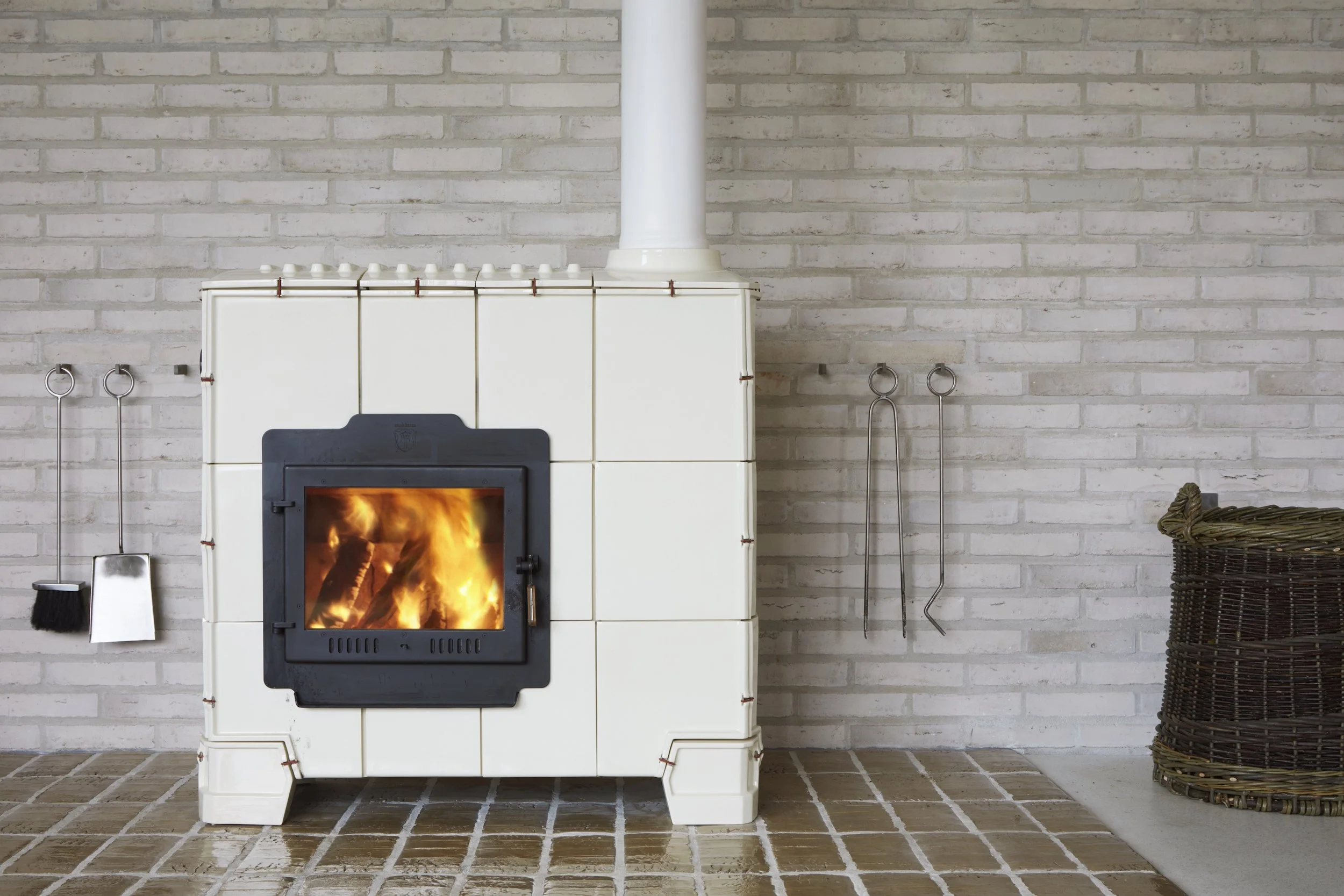 A fireplace with a burning fire in a room with white brick walls and tiled floor. Fireplace tools and a wicker basket are nearby.