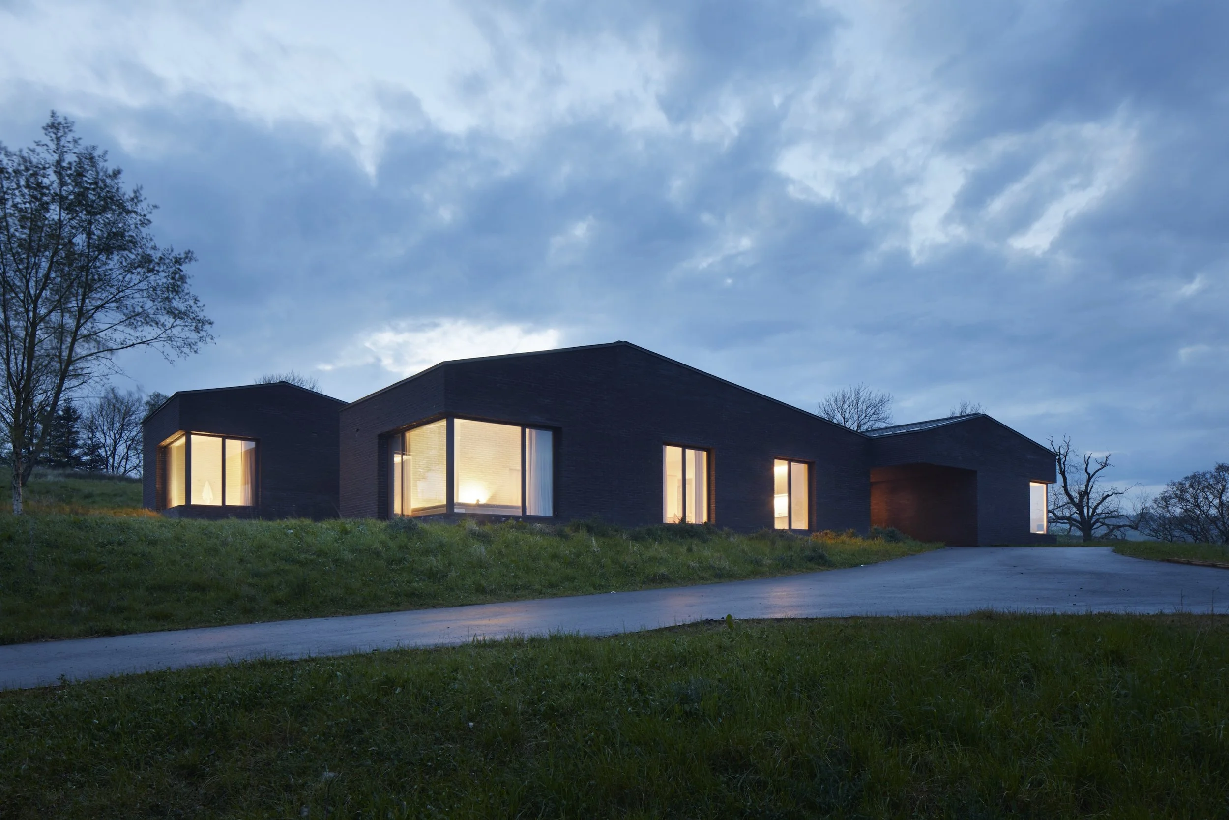 Modern black house with large windows illuminated inside, situated on a grassy hill under a cloudy evening sky.