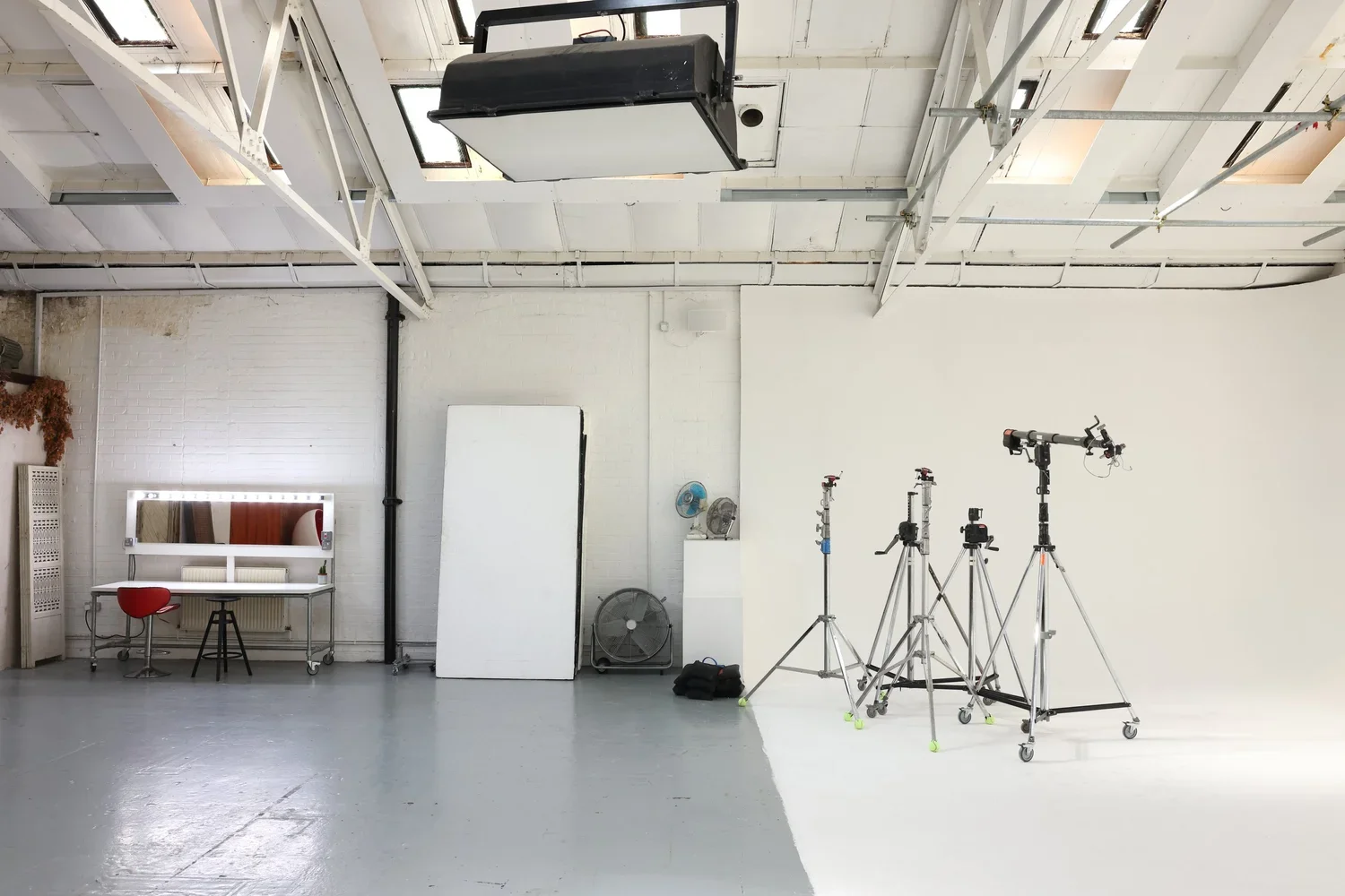 Empty photography studio with white walls, four tripods set up for filming, a white backdrop, ceiling skylights, and studio lighting equipment.