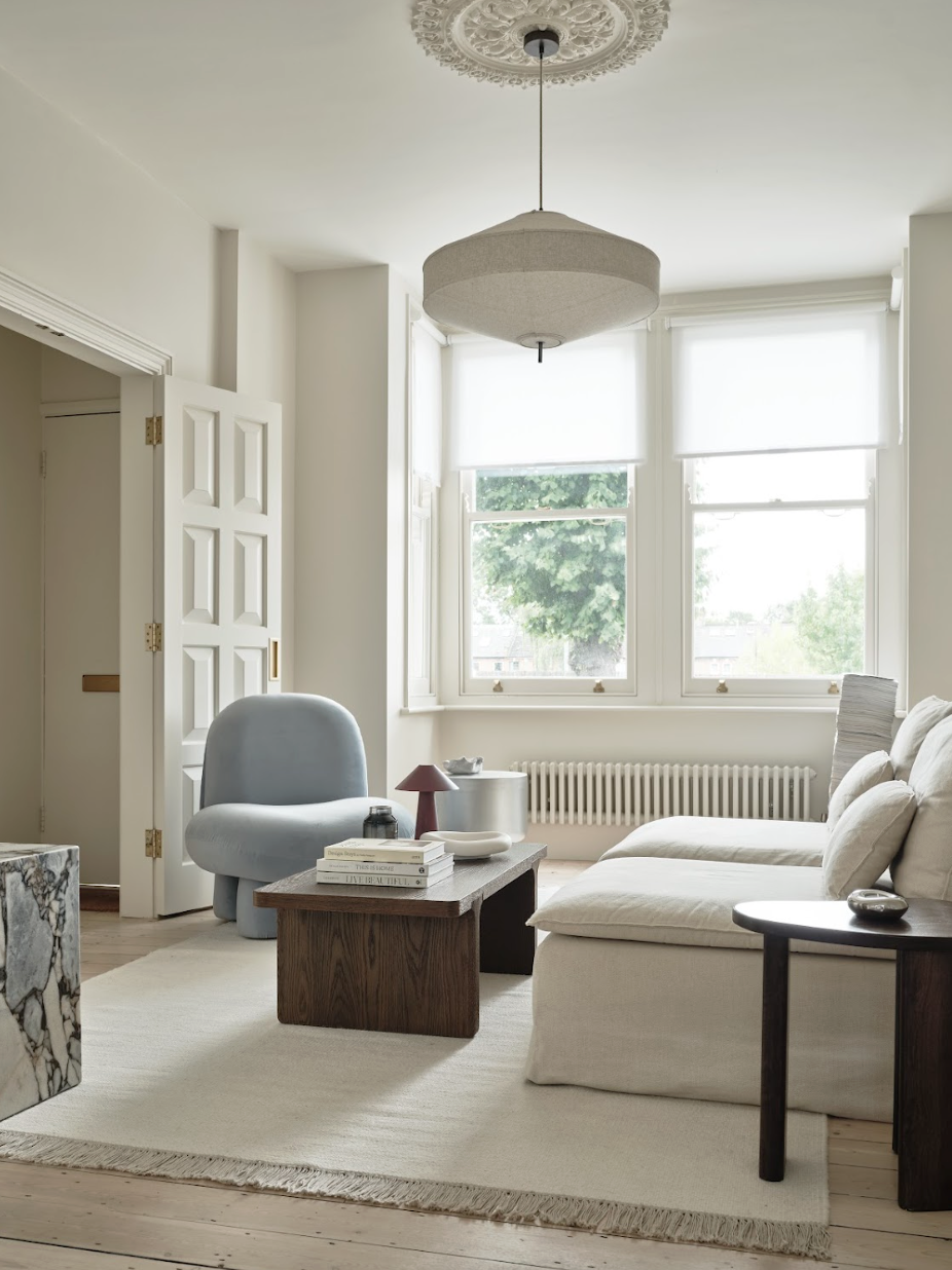 Bright living room with white walls, large windows with white blinds, beige sectional sofa, blue armchair, wooden coffee table, side tables, and a ceiling light fixture, with a view of green trees outside.