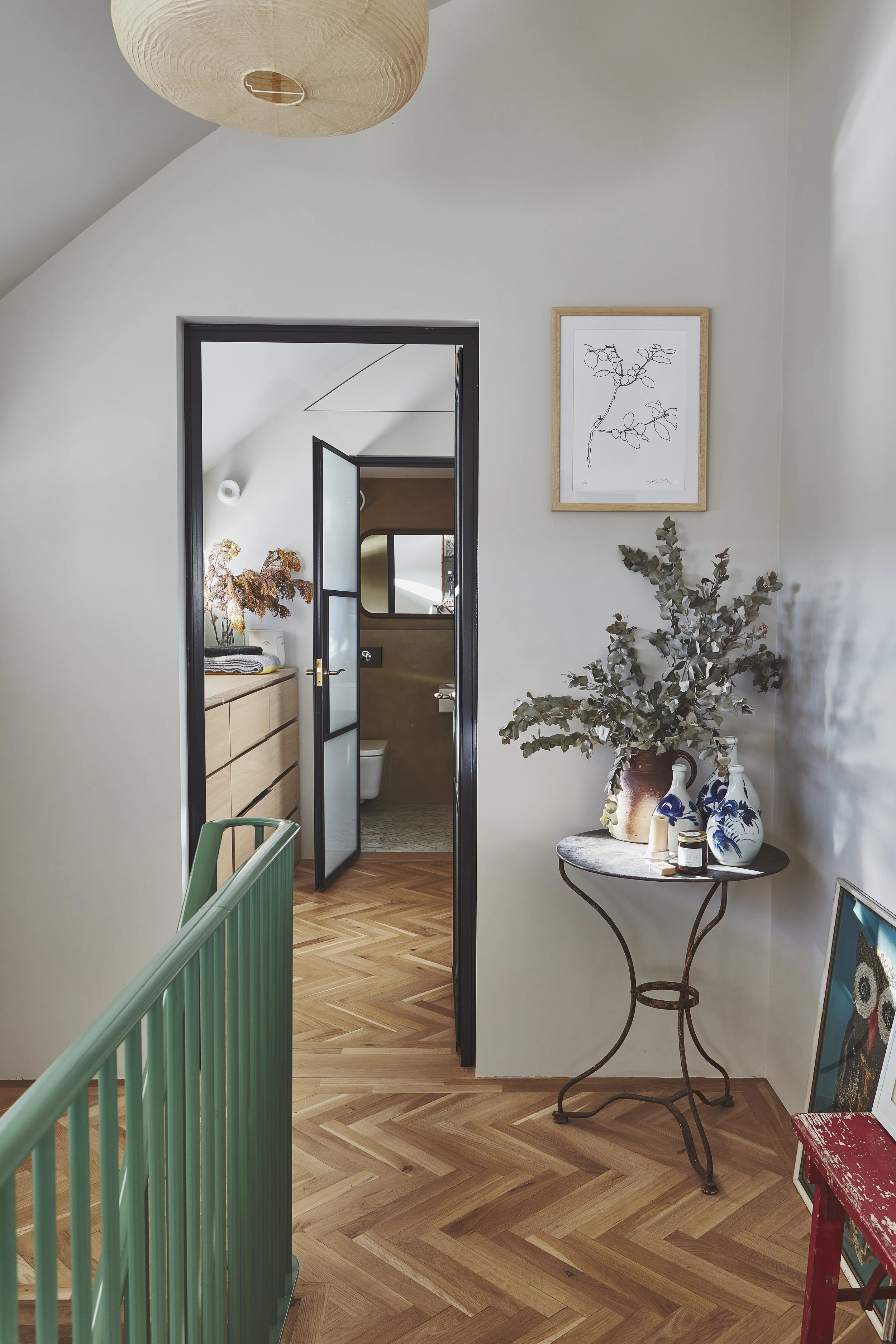 Interior view of a modern, cozy living space with a hallway leading to a bathroom. The space features a wooden herringbone floor, a black-framed glass door, a decorative table with vases and candles, a framed botanical print on the wall, and a large 