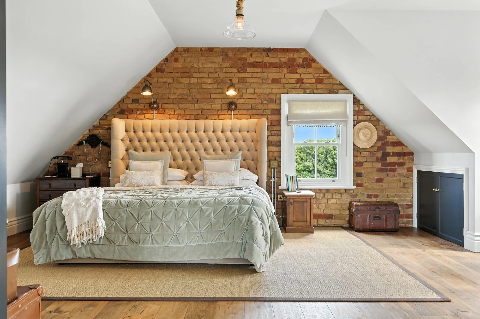A cozy bedroom with a brick accent wall behind the bed, featuring a tall, tufted beige headboard, two bedside tables, and a window showing green trees outside. The room has sloped white ceilings and warm lighting.