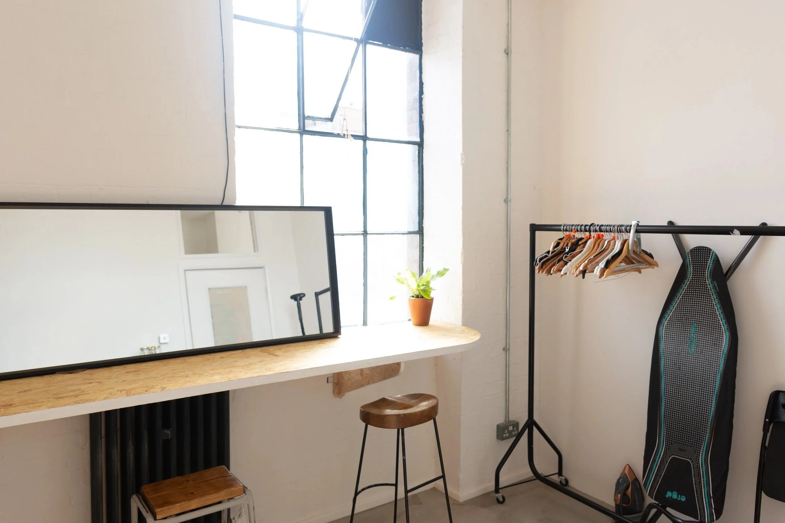Interior of a minimalistic room with a large window, a wooden counter with a potted plant and a mirror resting on it, a brown stool, a clothing rack with several hangers, and a standing electronic skateboard.