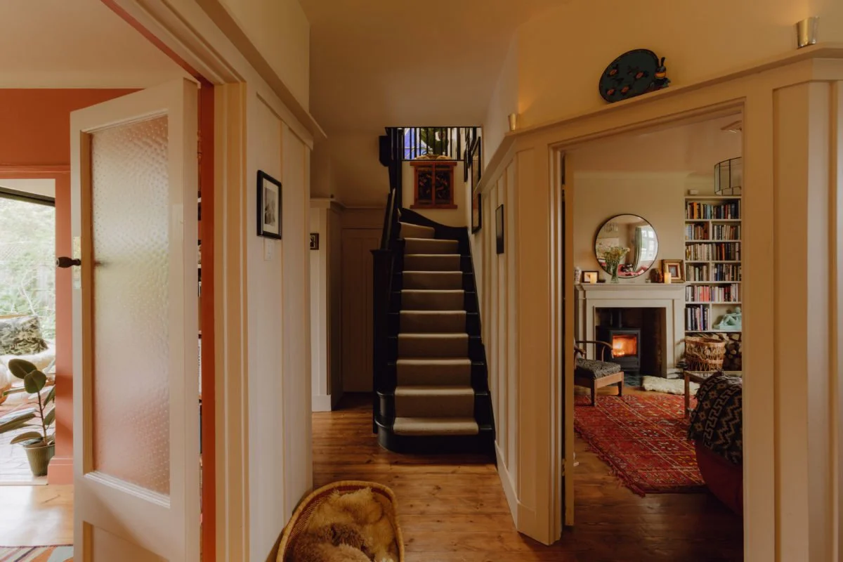 Indoor view of a home hallway with a staircase, leading to the upper floor, and a living room with a fireplace visible through an open doorway. The hallway has wooden flooring and minimal decorations.