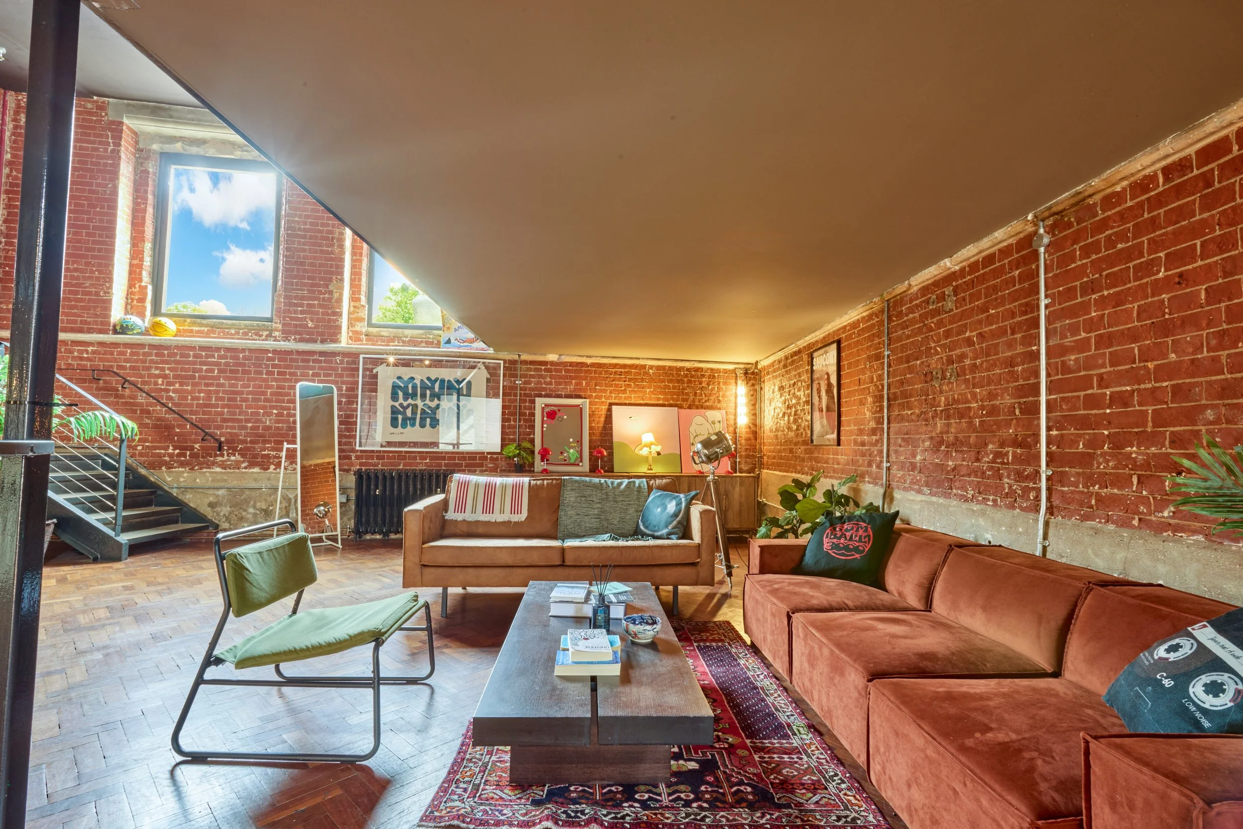 Living room with brick walls, large windows, a red velvet sofa, a beige sofa, a green chair, a coffee table with books, and various decorative items.