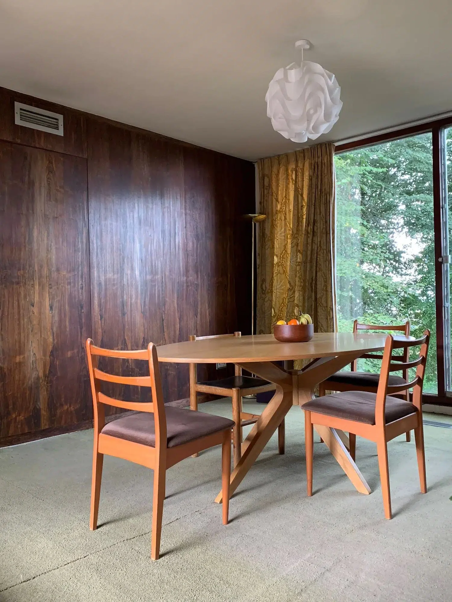 Dining room with wooden table and four chairs, large window with trees outside, brown wood-paneled wall, white ceiling with modern pendant light, and a bowl of fruit on the table.