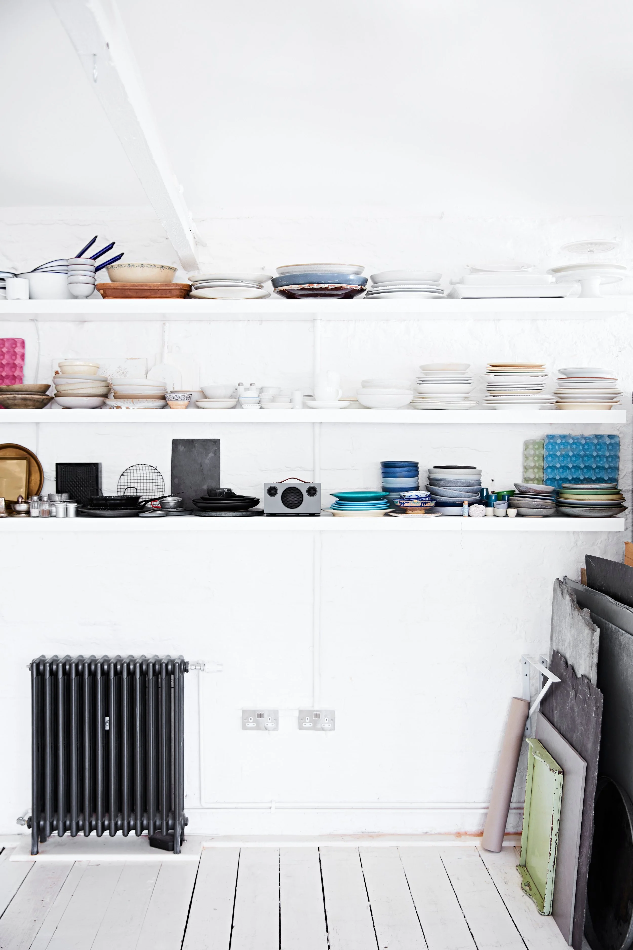 White shelving unit with stacked plates, bowls, and kitchenware, black radiator, and various items on the floor near a white wall.