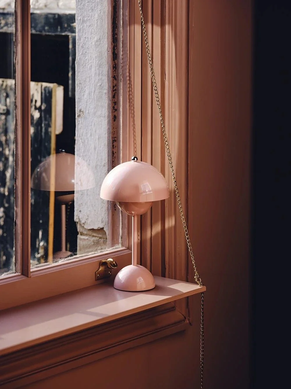 Pink table lamp with a curved shade on a windowsill next to a glass window reflecting the lamp and interior wood-paneled walls.