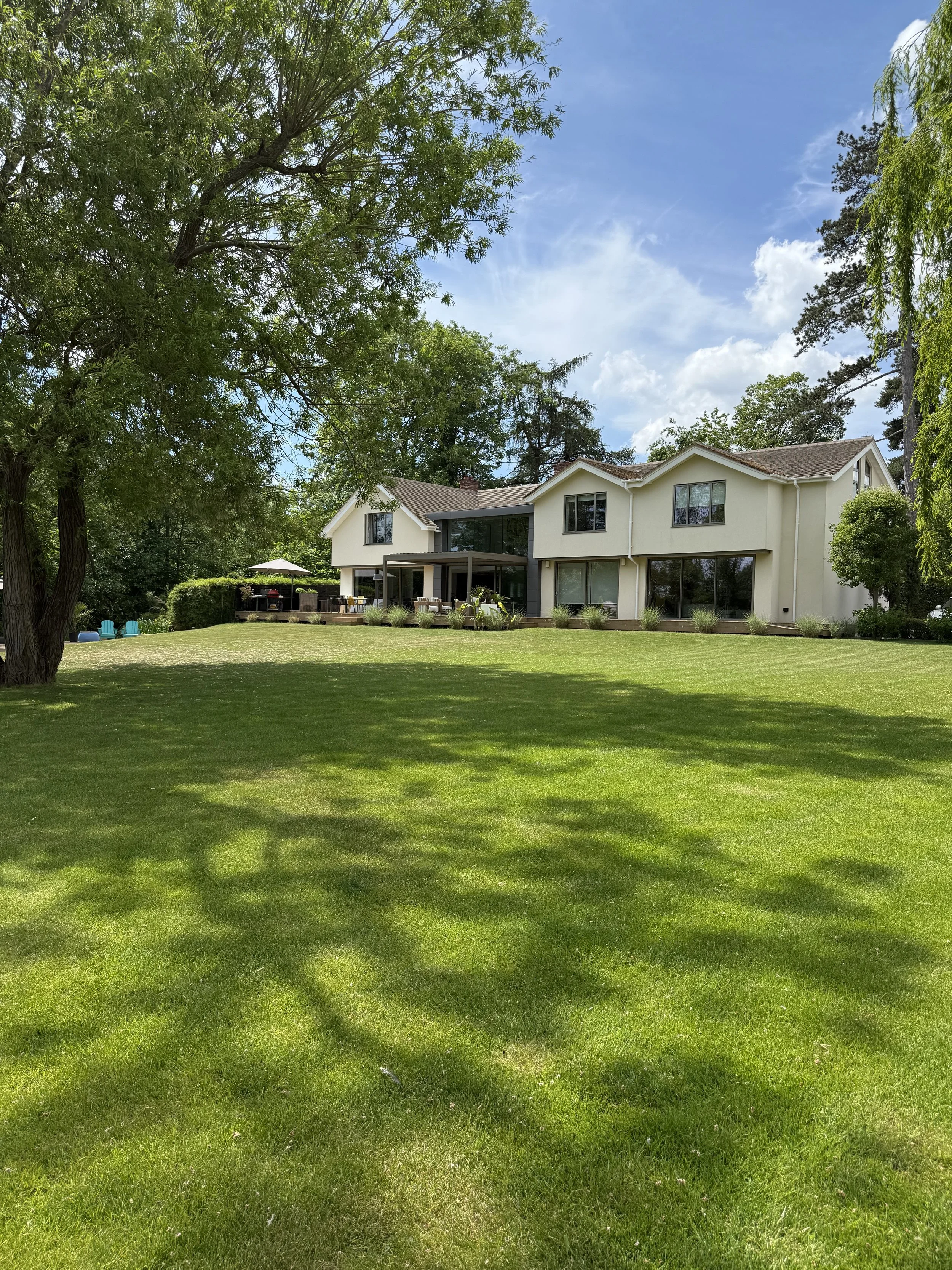 A large house with a white exterior and multiple windows, surrounded by green trees and a well-maintained grassy lawn on a sunny day with partly cloudy skies.
