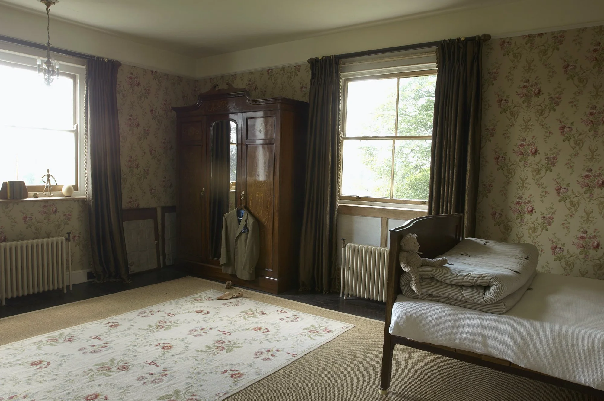 A bedroom with floral wallpaper, a single bed with a teddy bear and folded blanket, wooden wardrobe, two windows with curtains, a radiator, and a rug on the floor.