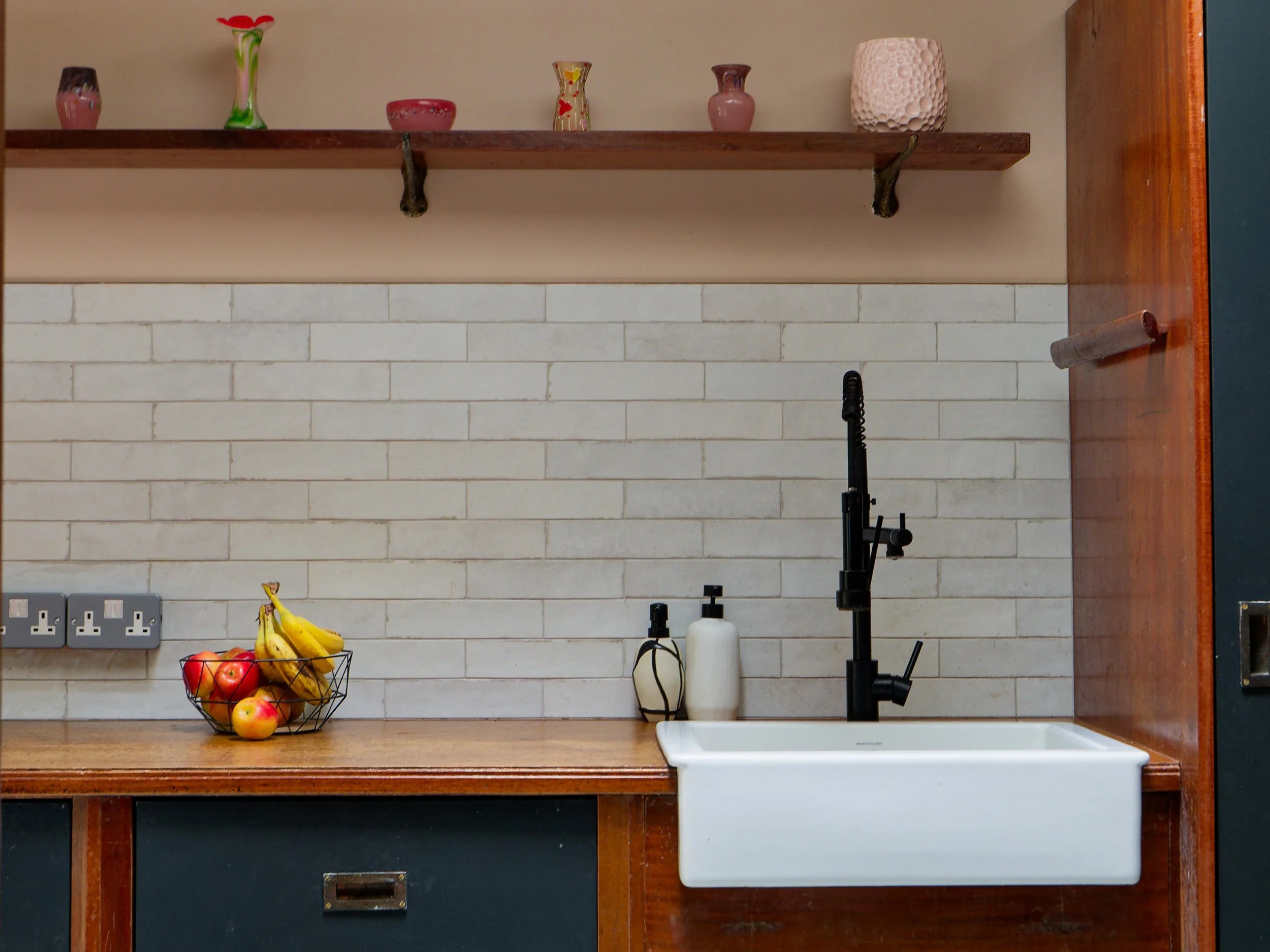 Kitchen with white brick backsplash, wooden countertop, and black faucet over a white farmhouse sink. A wire basket with bananas and apples on the counter, and soap dispensers nearby. Open wooden shelf with decorative vases and jars.