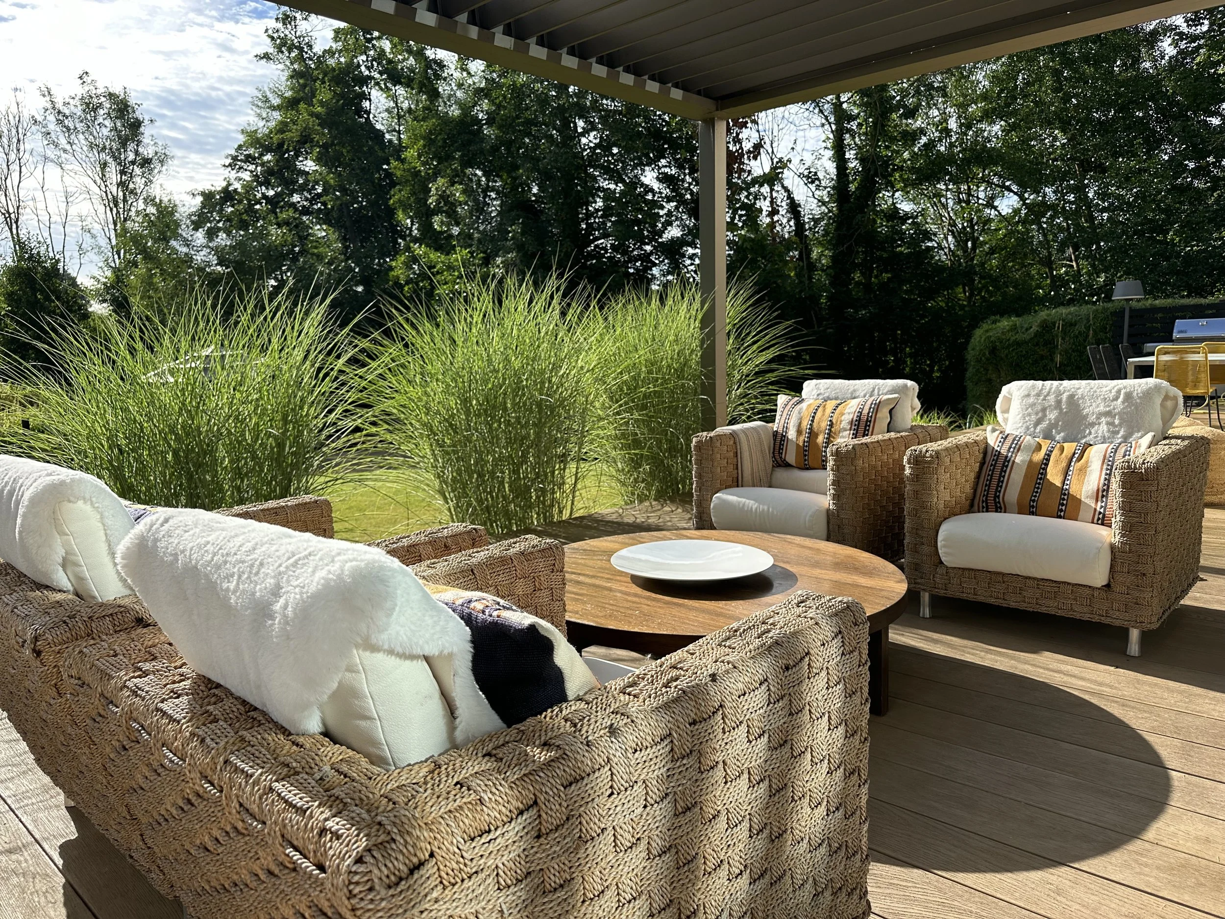 Outdoor patio seating area with wicker chairs, cushions, and a wooden table, overlooking lush greenery and trees.