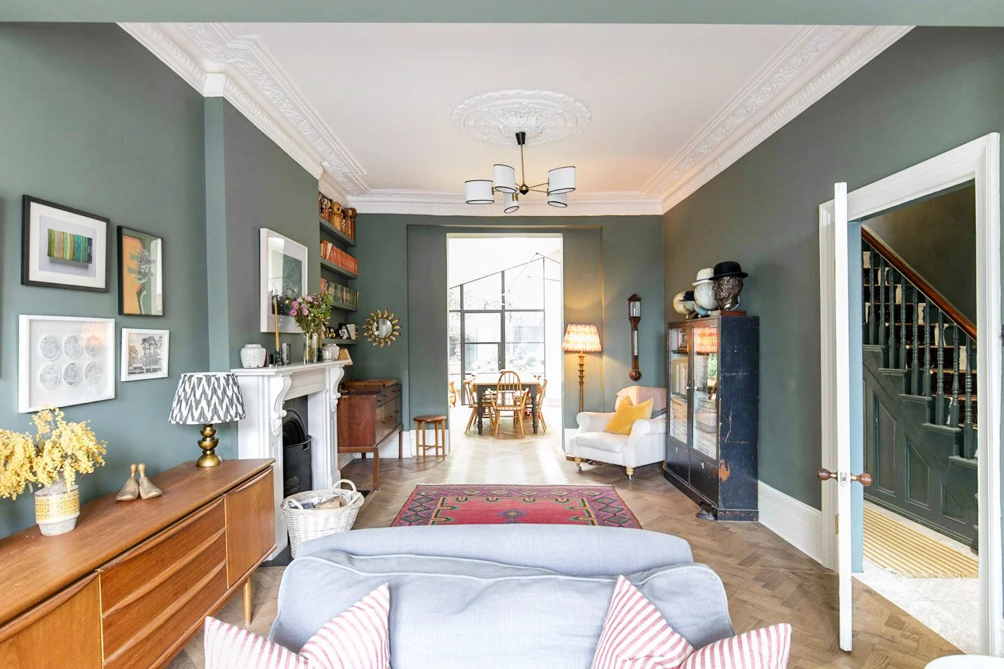 Living room interior with green walls, hardwood floors, vintage furniture, artwork, bookshelf, fireplace, and a dining area visible through an open doorway.