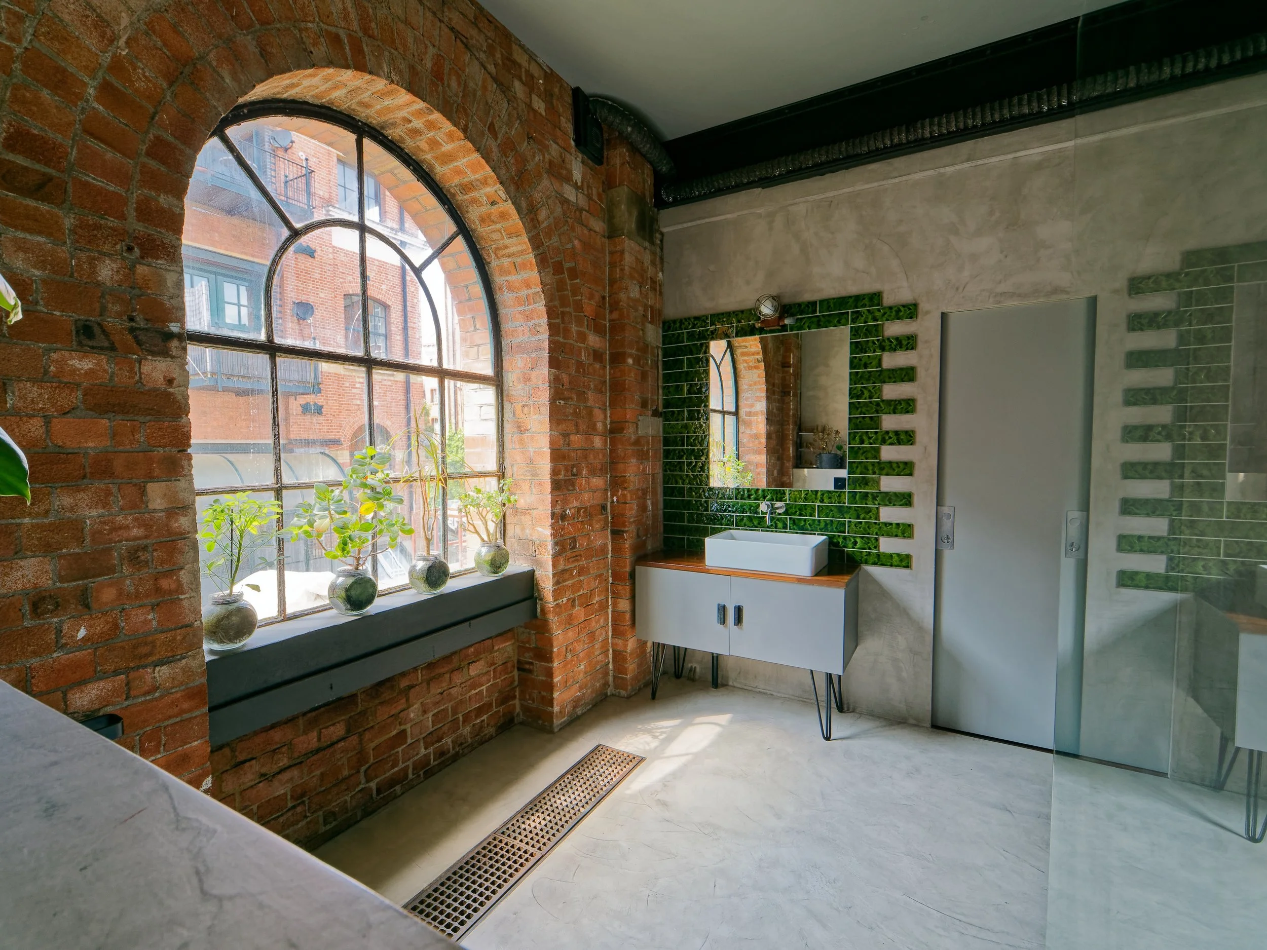 Modern bathroom with a large arched window, brick wall, green tile accents behind a white vanity, and a glass shower door.