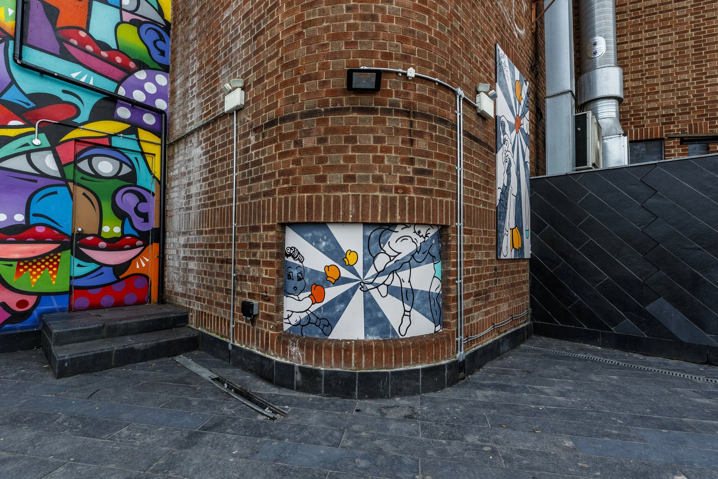 Colorful street art murals on brick building, featuring abstract face and cartoon characters.