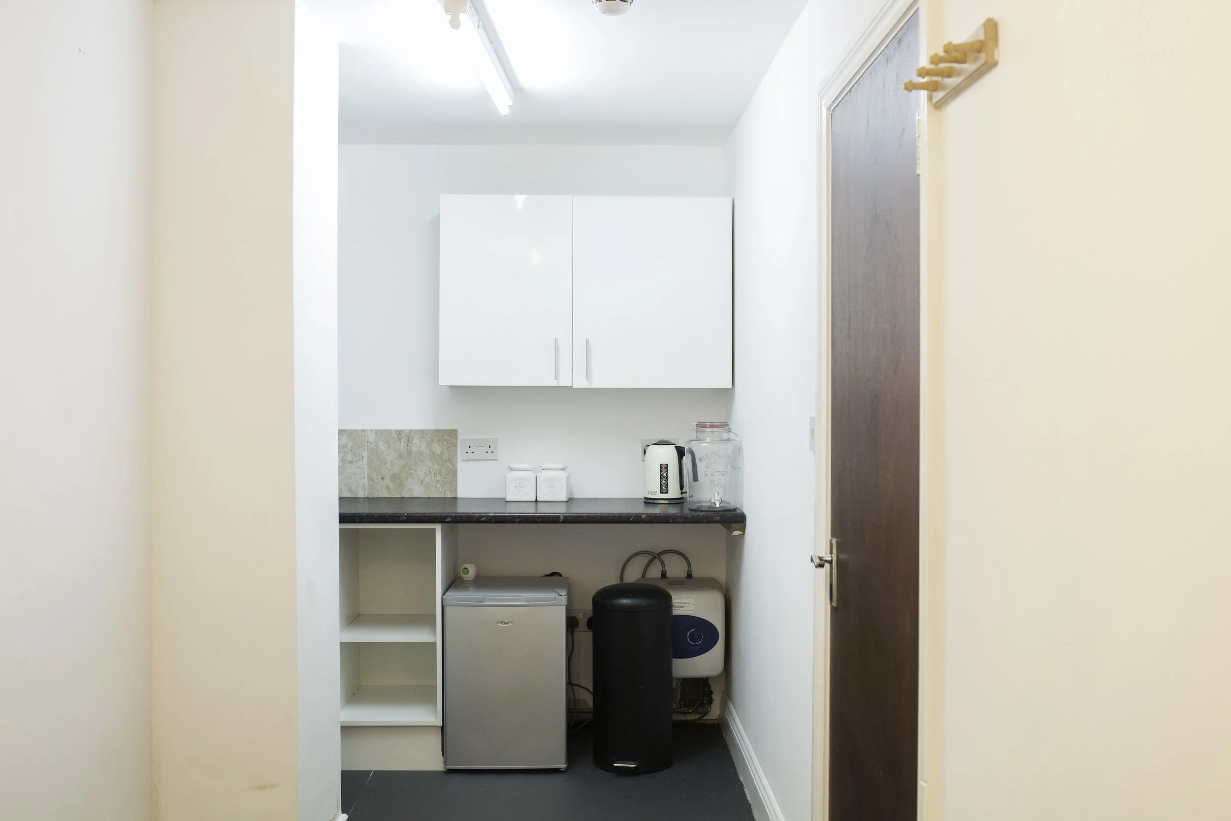 Small kitchen area with white cabinets, a black countertop, and appliances including a kettle, water dispenser, mini fridge, and black water filter unit.