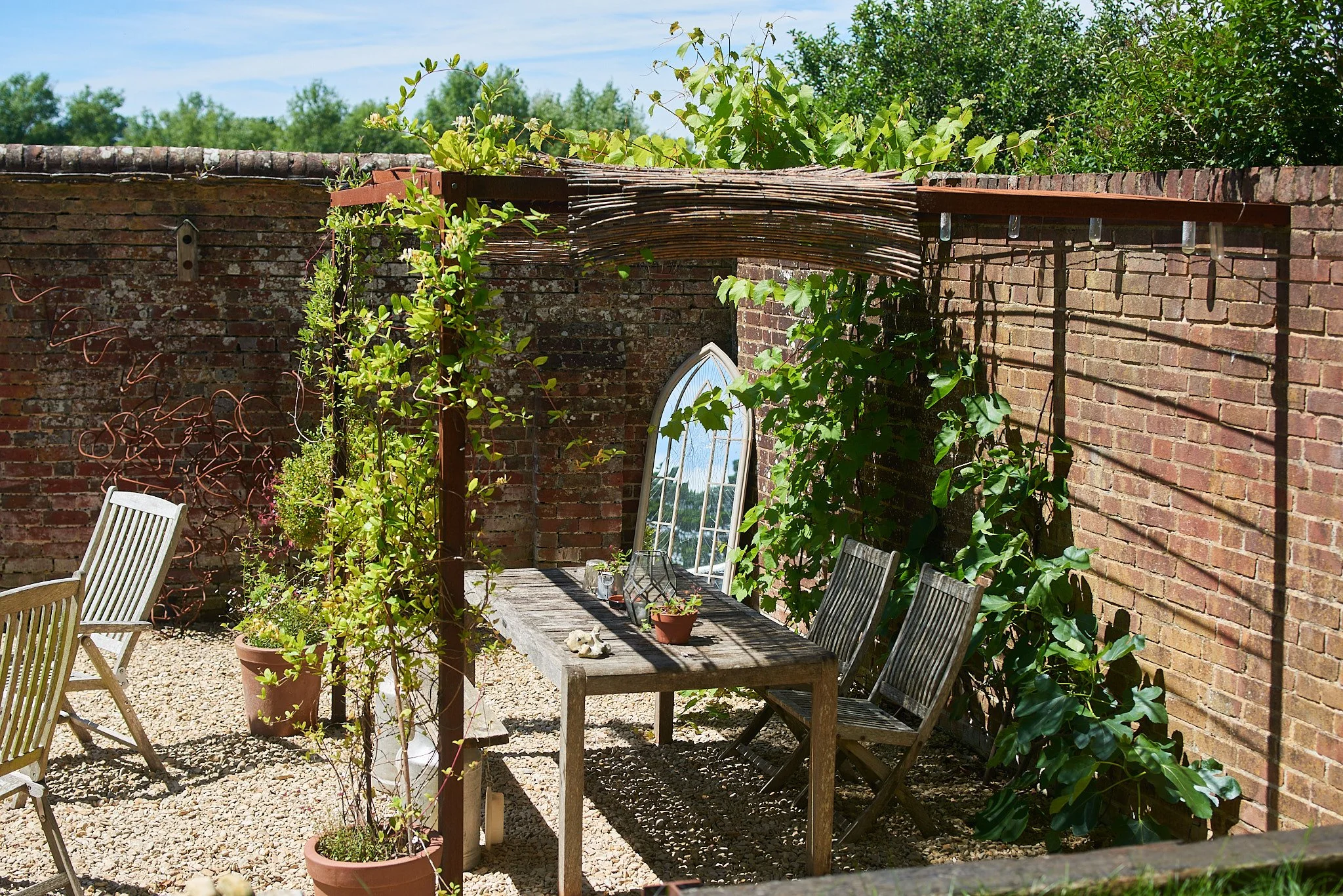 A small backyard patio with a rustic wooden table and two matching chairs, surrounded by potted plants, climbing vines, and a tall mirror leaning against a brick wall, under a sunny sky.