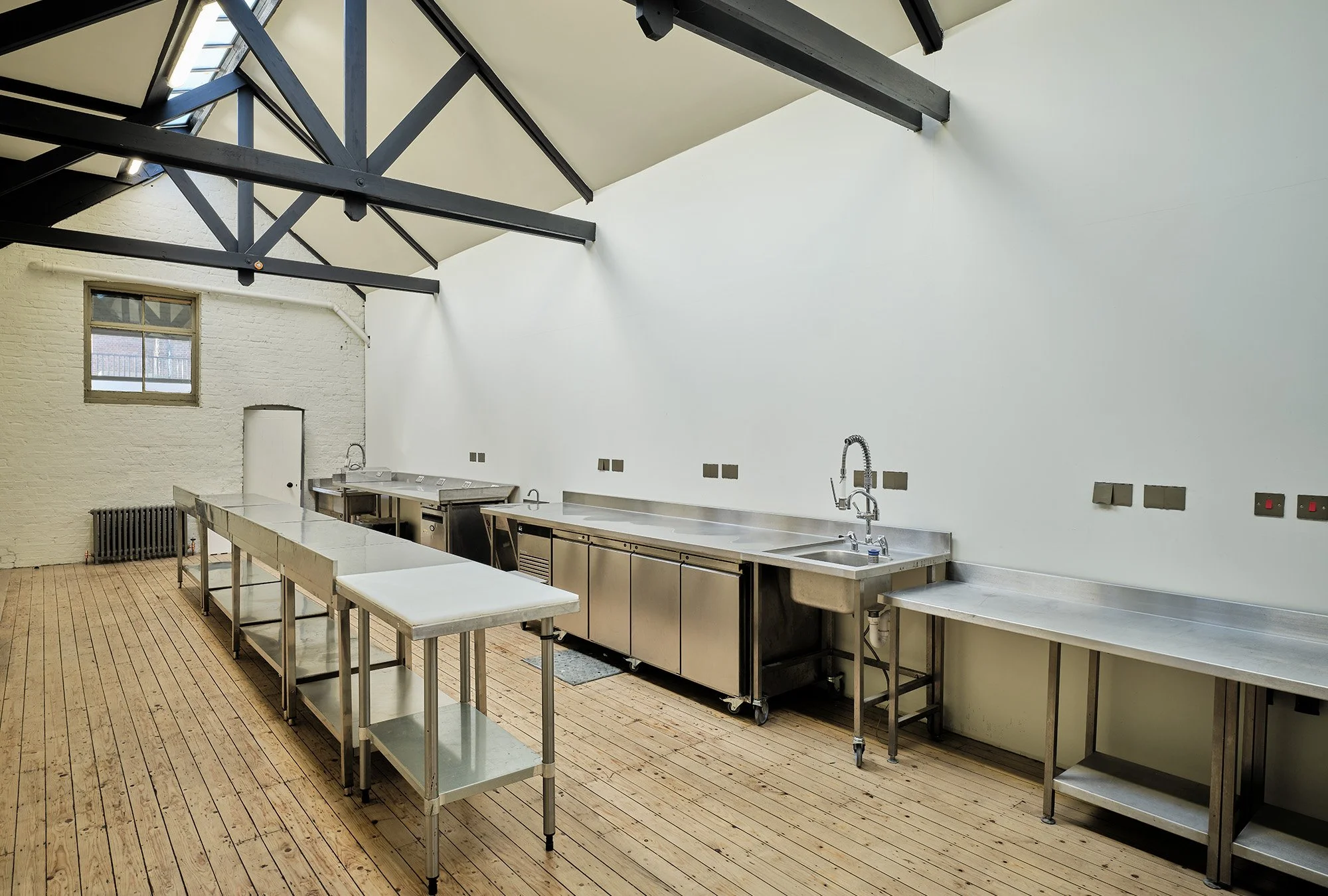 A clean commercial kitchen with stainless steel counters and appliances, wooden plank floor, and a large window letting in natural light.