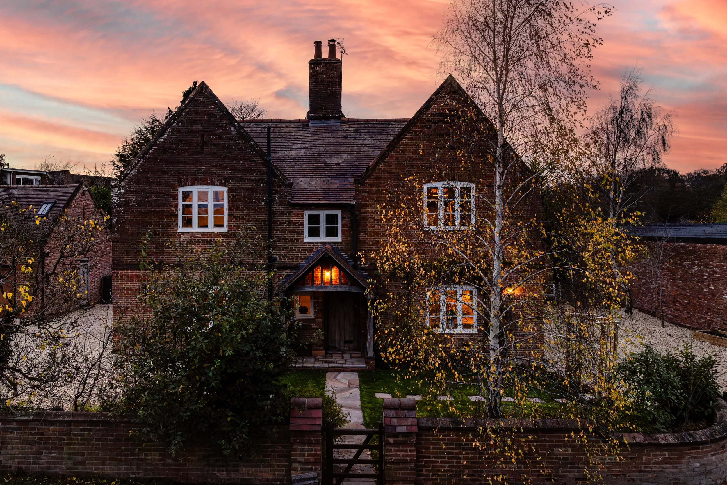 A brick house with lit windows during sunset, surrounded by trees and a brick fence.