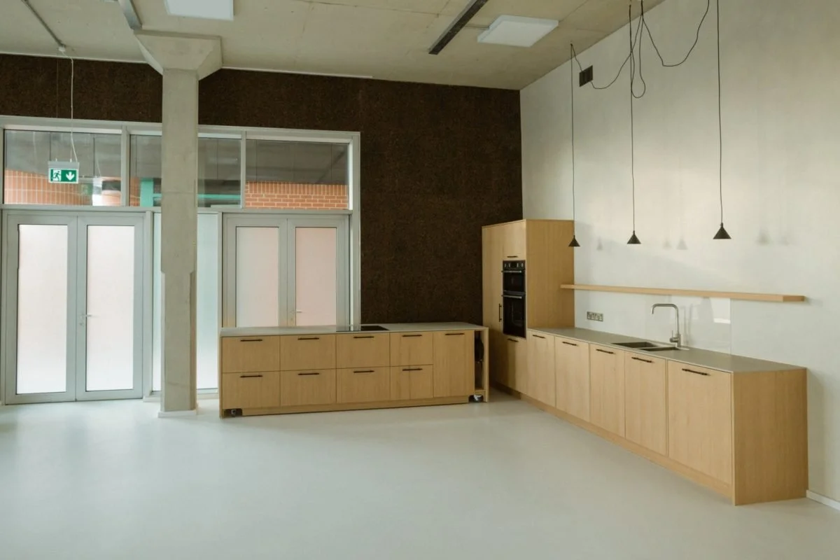 Empty kitchen with light wooden cabinets, black handles, and modern black hanging pendant lights, with a built-in oven and stove, a sink, and a wooden shelf on the white wall.