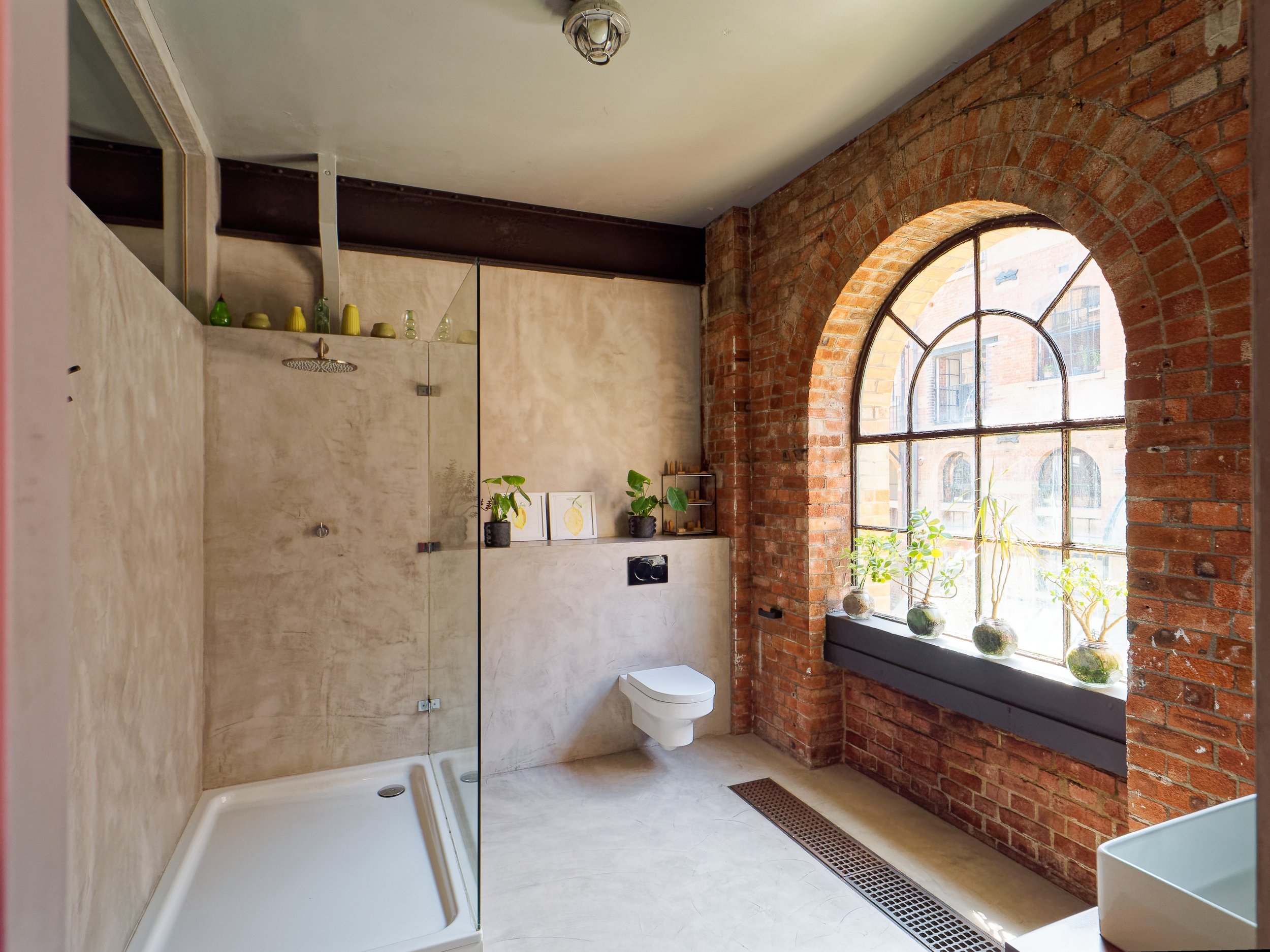 Bathroom with exposed brick wall, large arched window with plants on the window sill, walk-in shower with a glass partition, and a floating toilet.