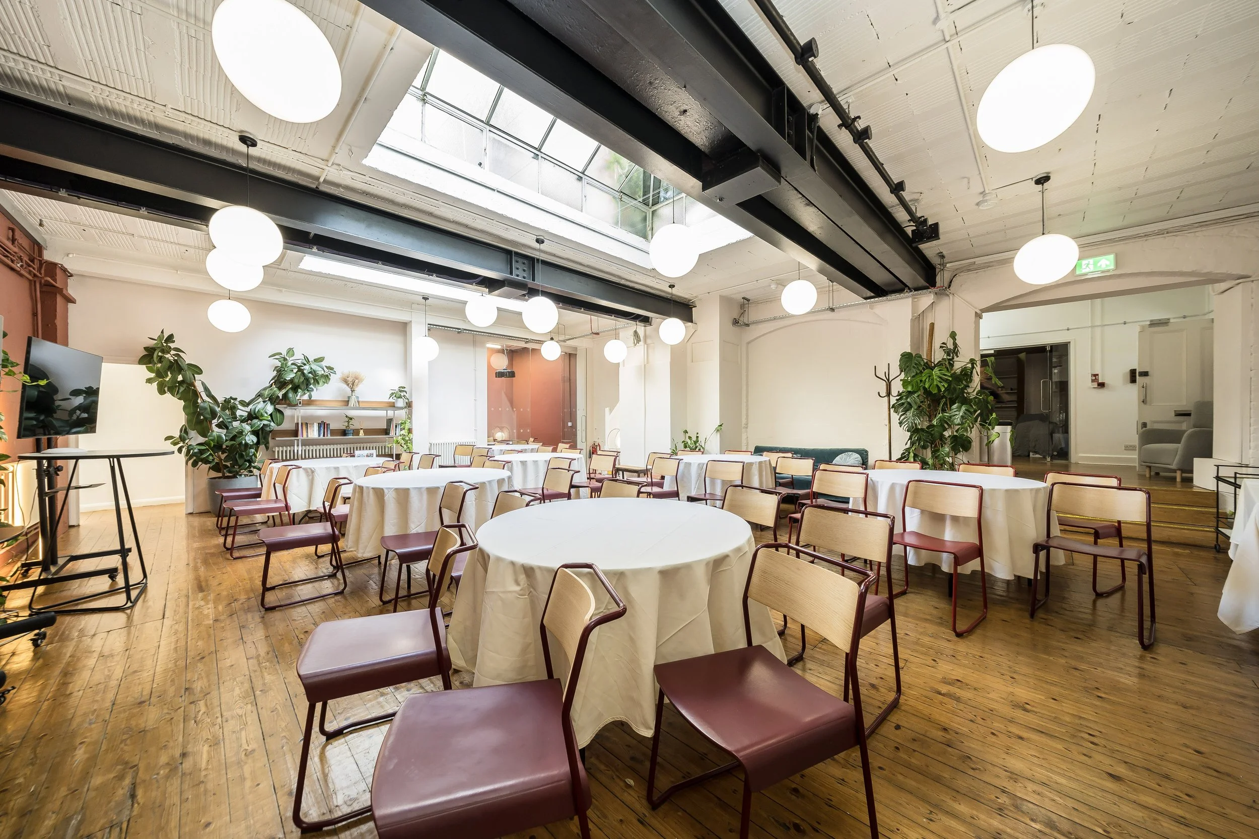 An empty event space with round tables covered with white tablecloths and red chairs on a wooden floor. Bright lighting from ceiling fixtures and a skylight, with indoor plants and a TV on the left.