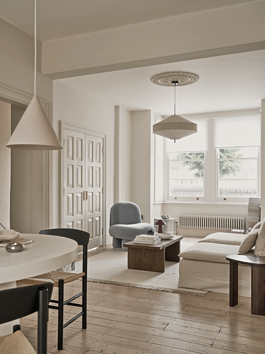 Living room with beige walls, large window with white blinds, white sectional sofa, gray armchair, wooden side table, round white dining table, and minimalist pendant lighting.