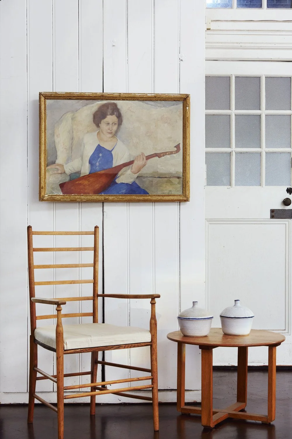 A painting of a woman holding a musical instrument, hanging on a white paneled wall. Below the painting, a wooden chair with a white cushion and a small wooden table with two ceramic jars are seen.