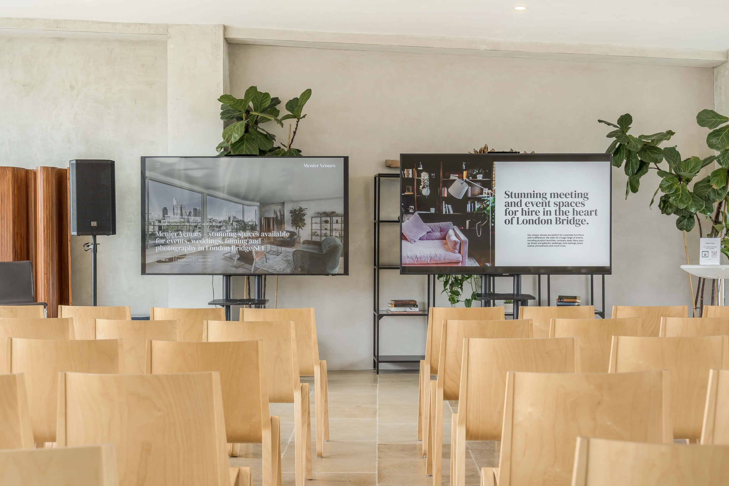 A conference room set up with chairs, two large screens displaying promotional content for venue spaces, surrounded by indoor plants, with a white wall background.