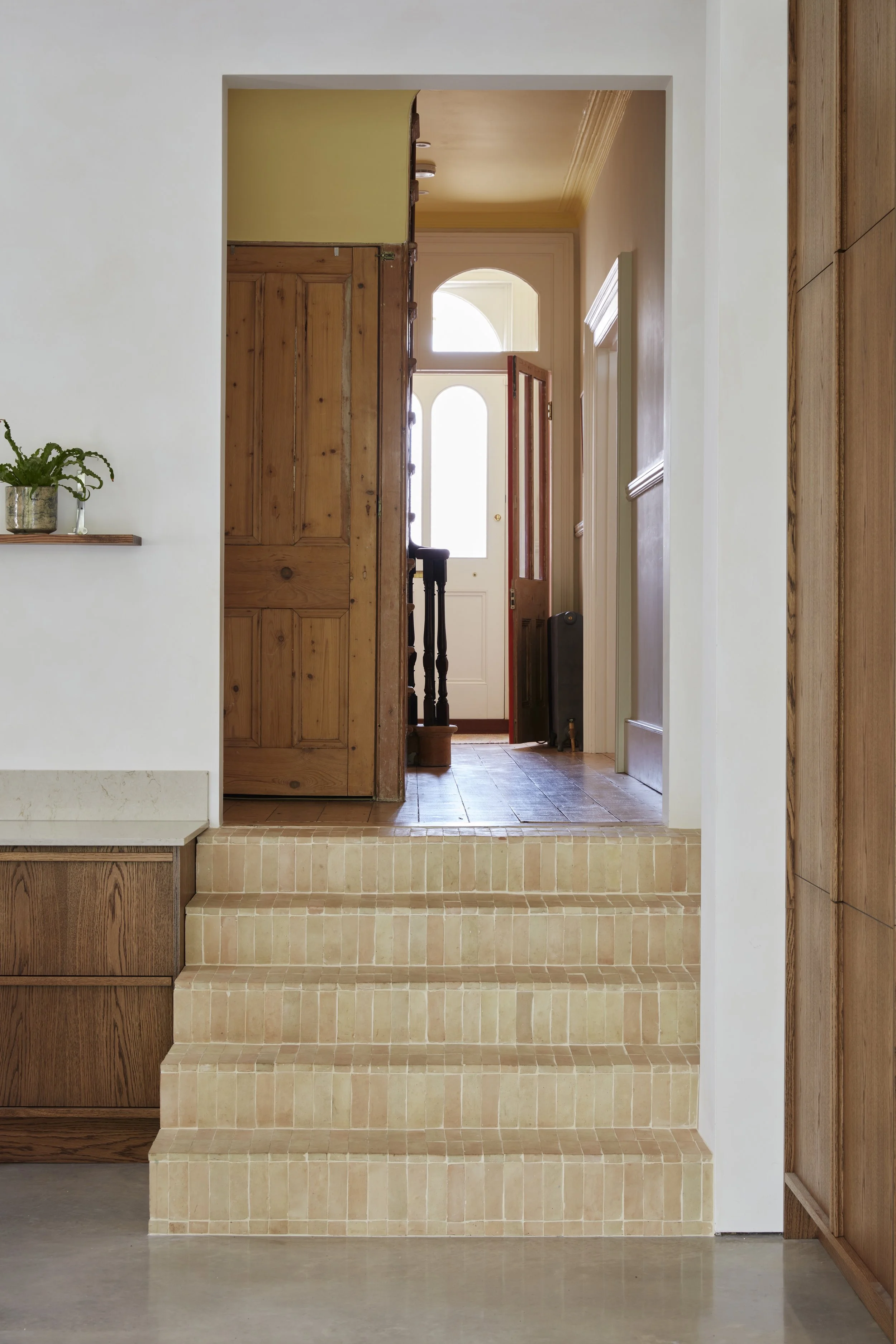 Interior view of a home entrance with tiled steps leading to a wooden floor and a door with glass panels, surrounded by wooden and white walls.
