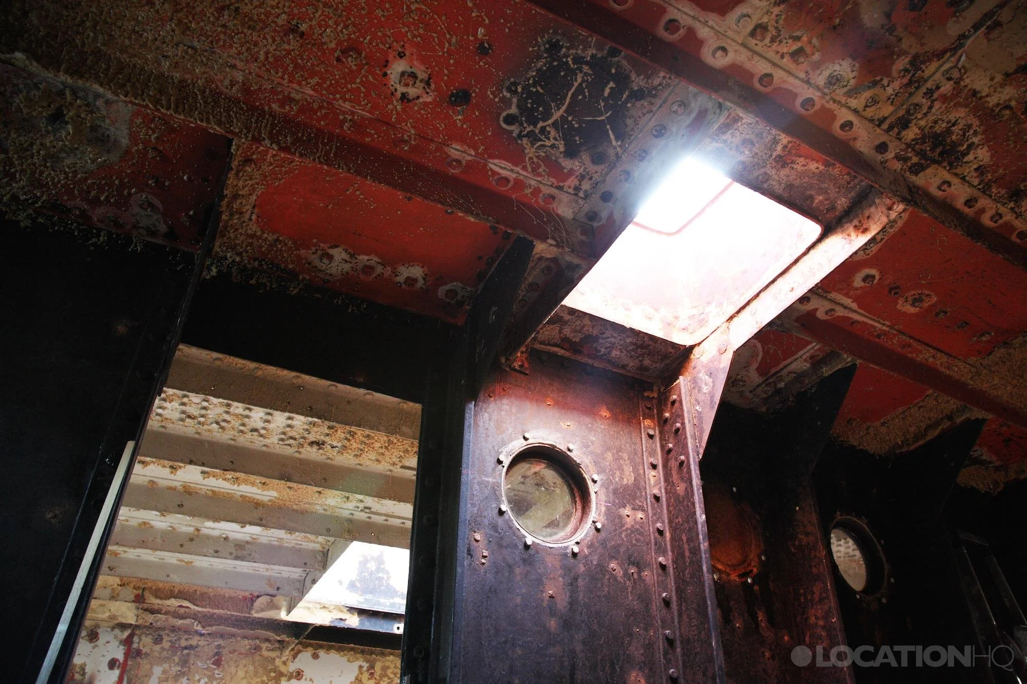 Interior of an old, rusted, and weathered ship with metal walls and ceiling, showing signs of corrosion and peeling paint. A small circular port hole is visible on the wall, with sunlight coming through a rectangular opening on the ceiling, illuminat