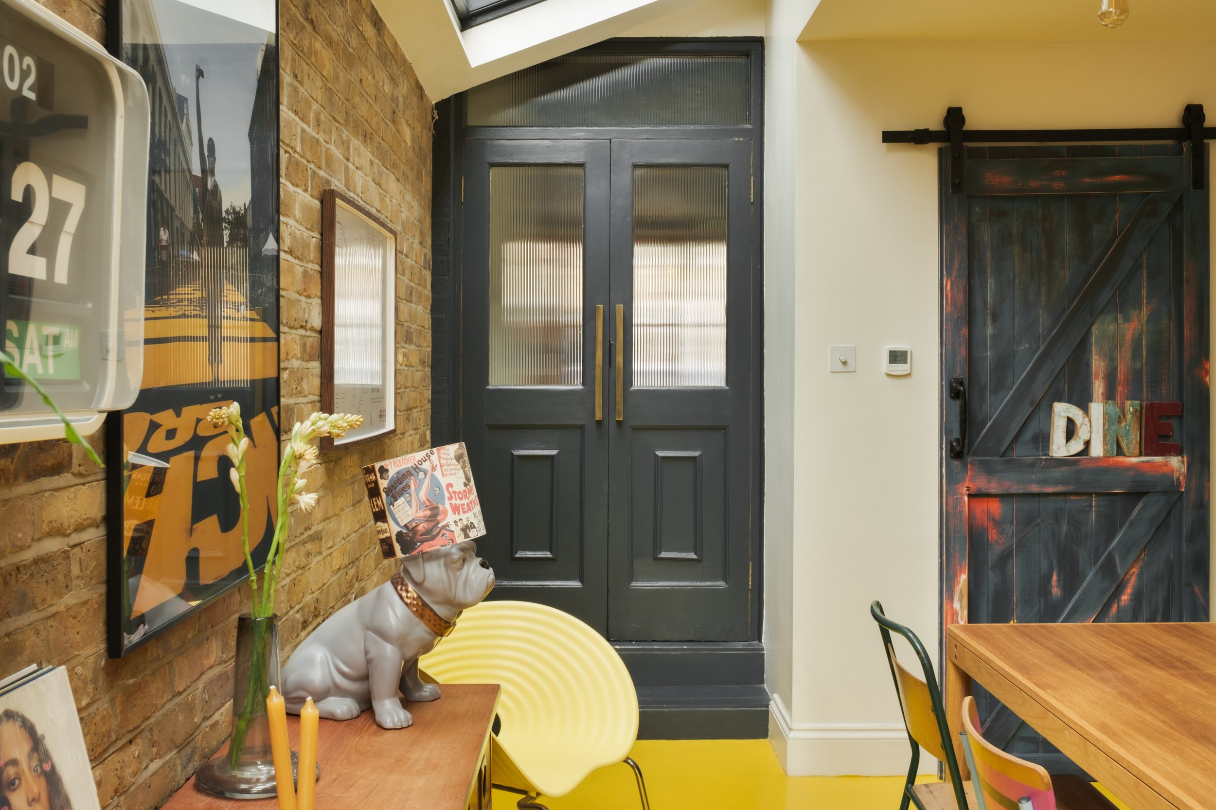 Interior of a room with a brick wall and various decor items, including a ceramic dog statue with a magazine on its head, a yellow chair, a wooden table, a dark barn door with the word 'DINE' on it, and glass double doors.