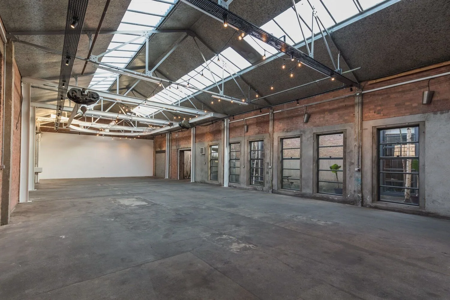 Empty industrial-style room with large windows, exposed brick walls, and overhead lighting, featuring a high ceiling with skylights and metal beams.