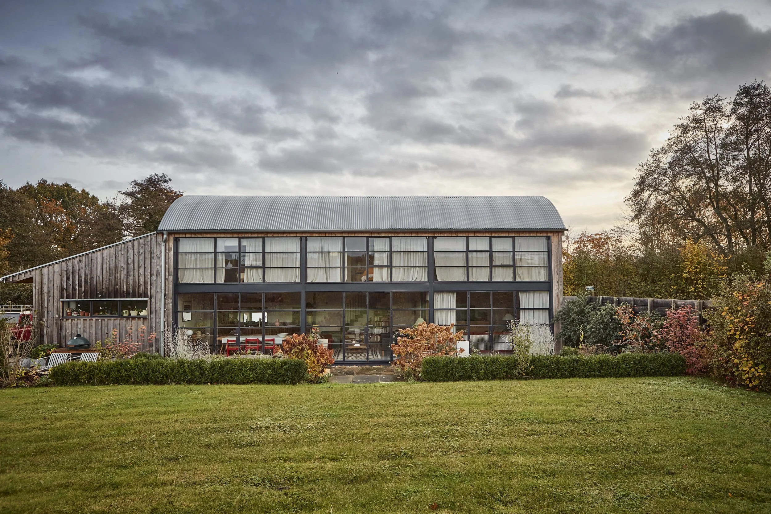 A modern two-story house with large windows and a peaked corrugated metal roof, surrounded by a lush garden with colorful plants and trees, under a cloudy sky.