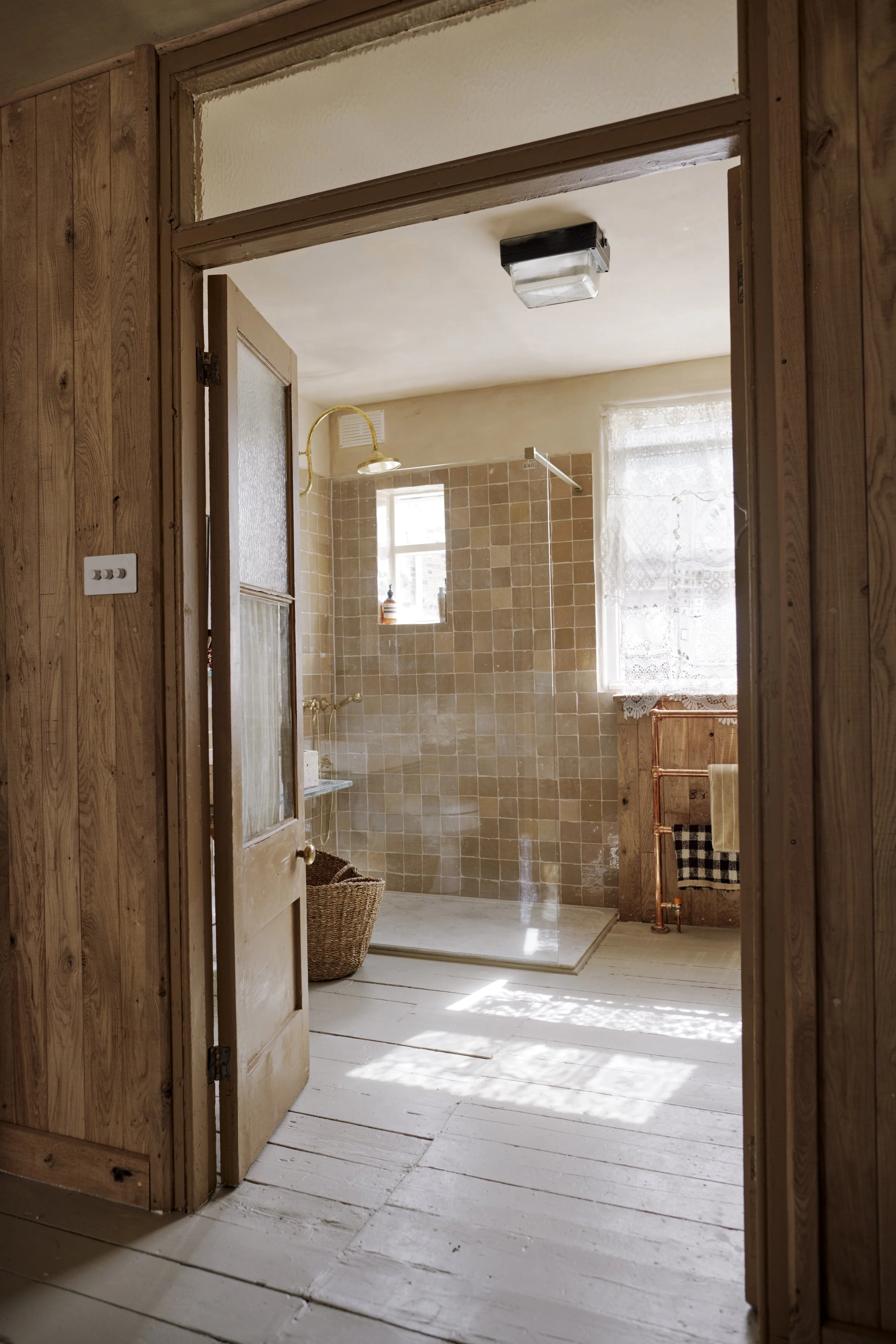 View into a rustic bathroom with a walk-in shower, beige tile walls, a small window, a copper pipe towel rack, and a wire basket. Sunlight filters through a lacy curtain on the larger window.