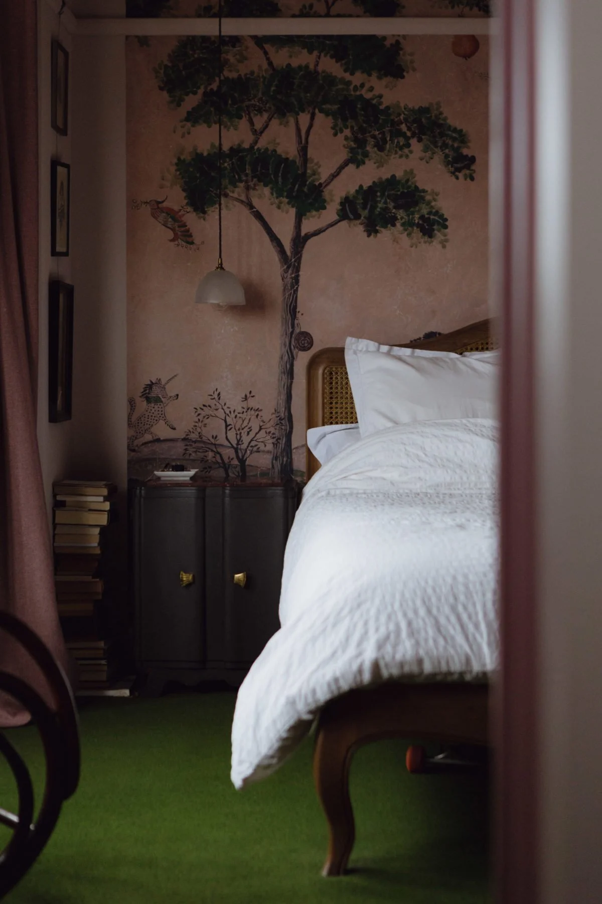 A cozy bedroom with a large white bed, artwork featuring a tree on the wall, a dark bedside cabinet with brass handles, and a green carpet.